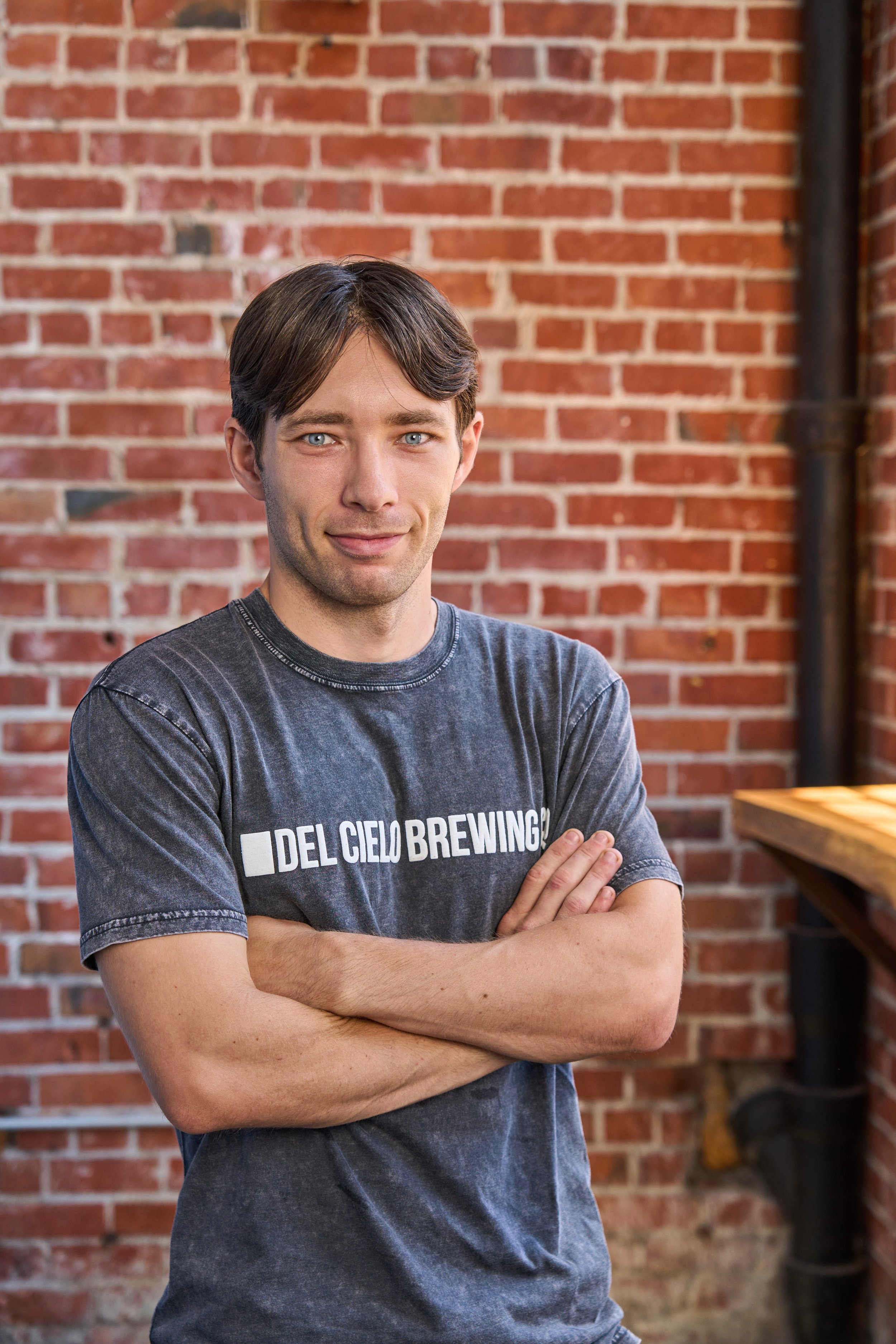 A man with dark brown hair and blue eyes crossing his arms, wearing a gray T-shirt with white text that reads 'DEL CIELO BREWING', standing in front of a red brick wall with some green plants hanging on the sides.