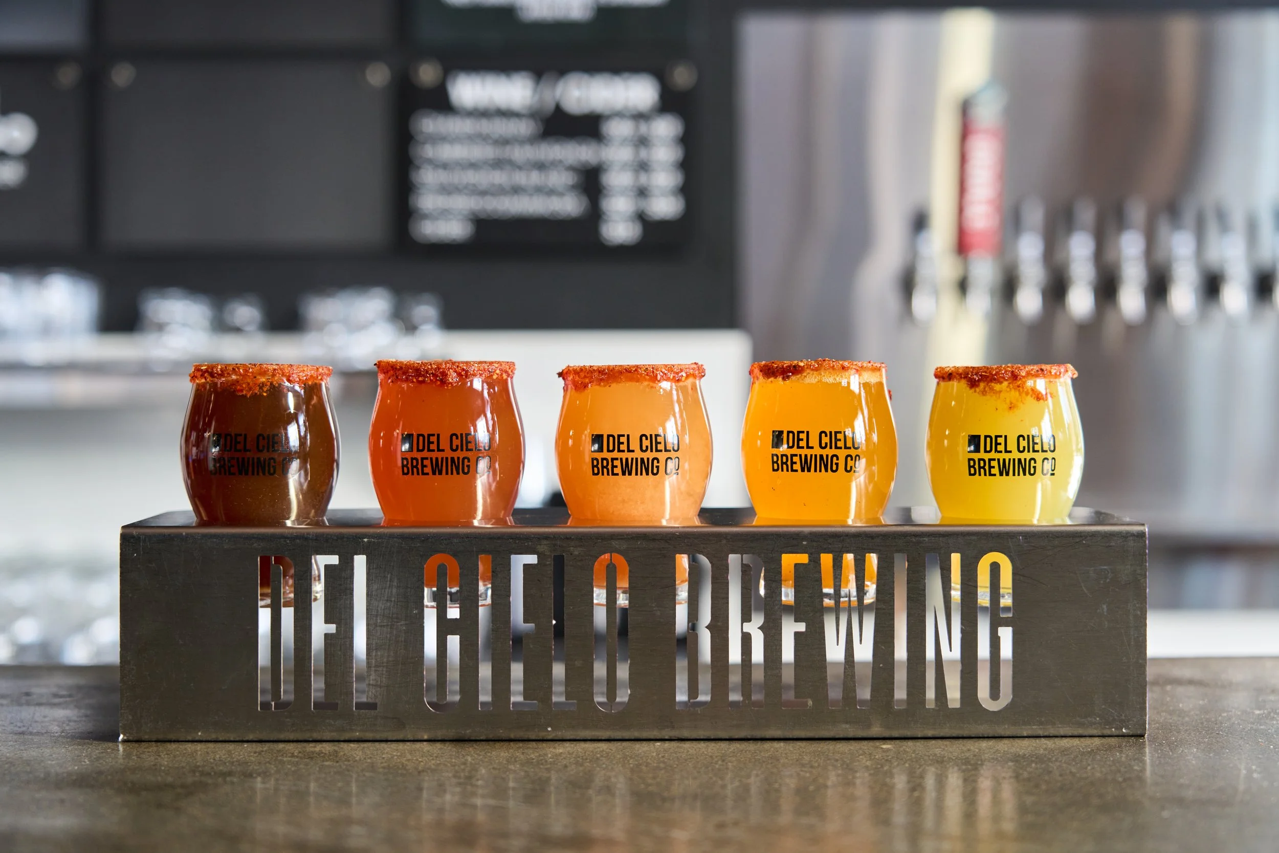 A row of five beer glasses filled with different shades of beer, from dark to light, on a metal beer flight tray with the logo and name of Del Cielo Brewing Co.