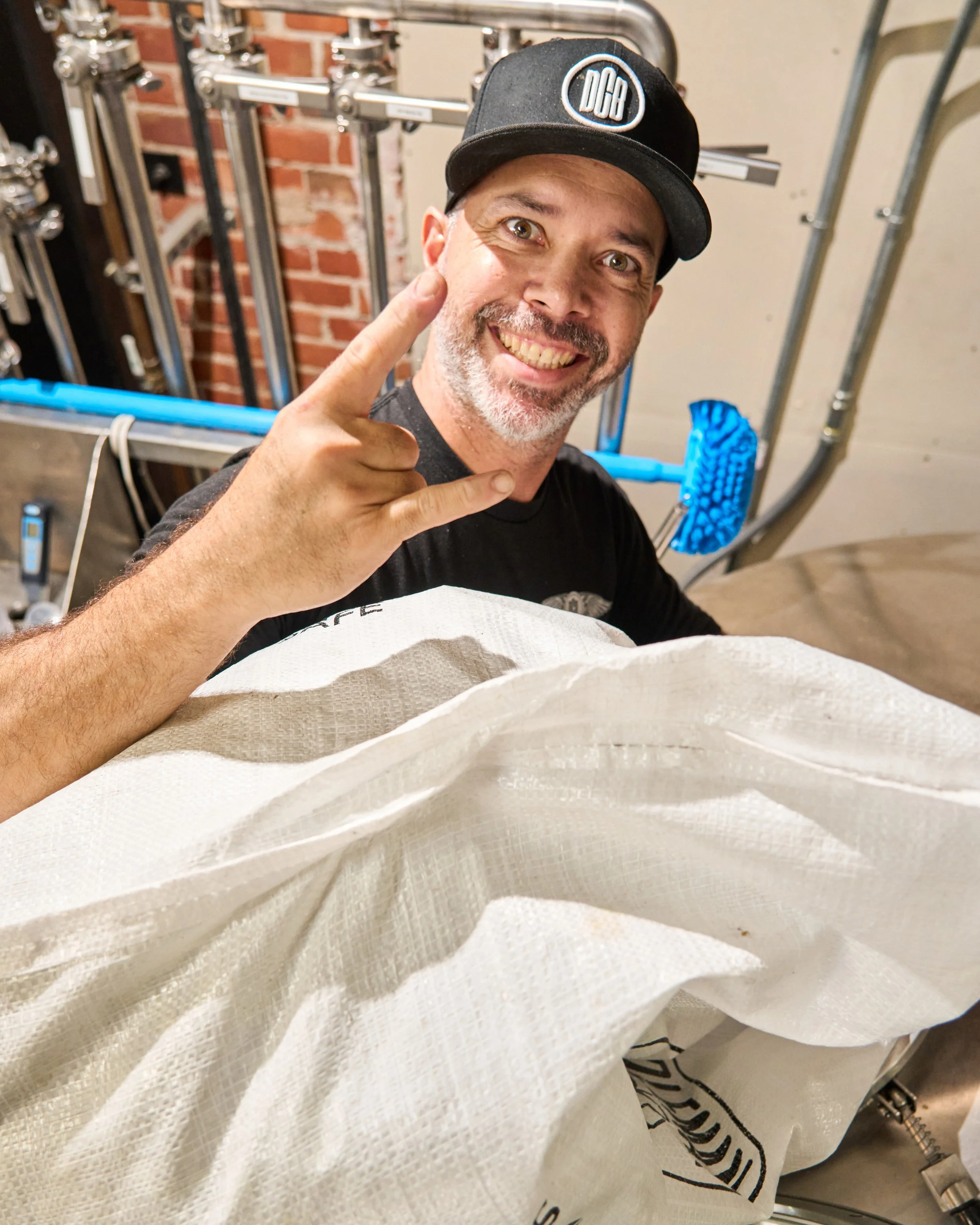 A man with a beard and a black cap with the logo 'DCB' is smiling and making a rock 'n' roll gesture with his hand. 