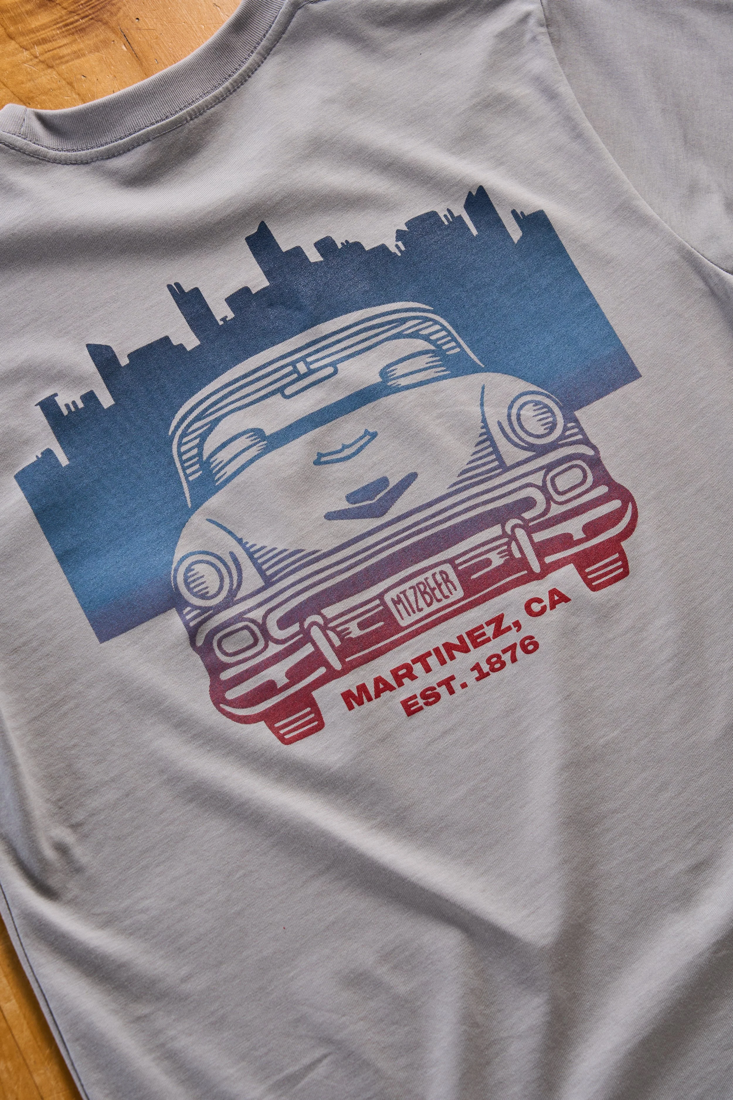 A gray t-shirt with a graphic of a vintage car and a cityscape in blue and red colors, featuring text about Martinez, California, established in 1876.