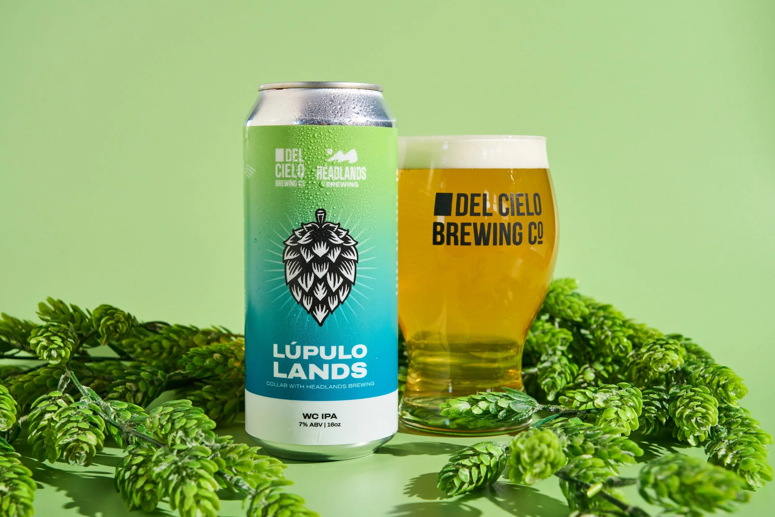 A can of Del Cielo Brewing Co.'s Lupulo Lands WC IPA with 7% ABV placed next to a glass of beer, surrounded by green hop cones against a light green background.