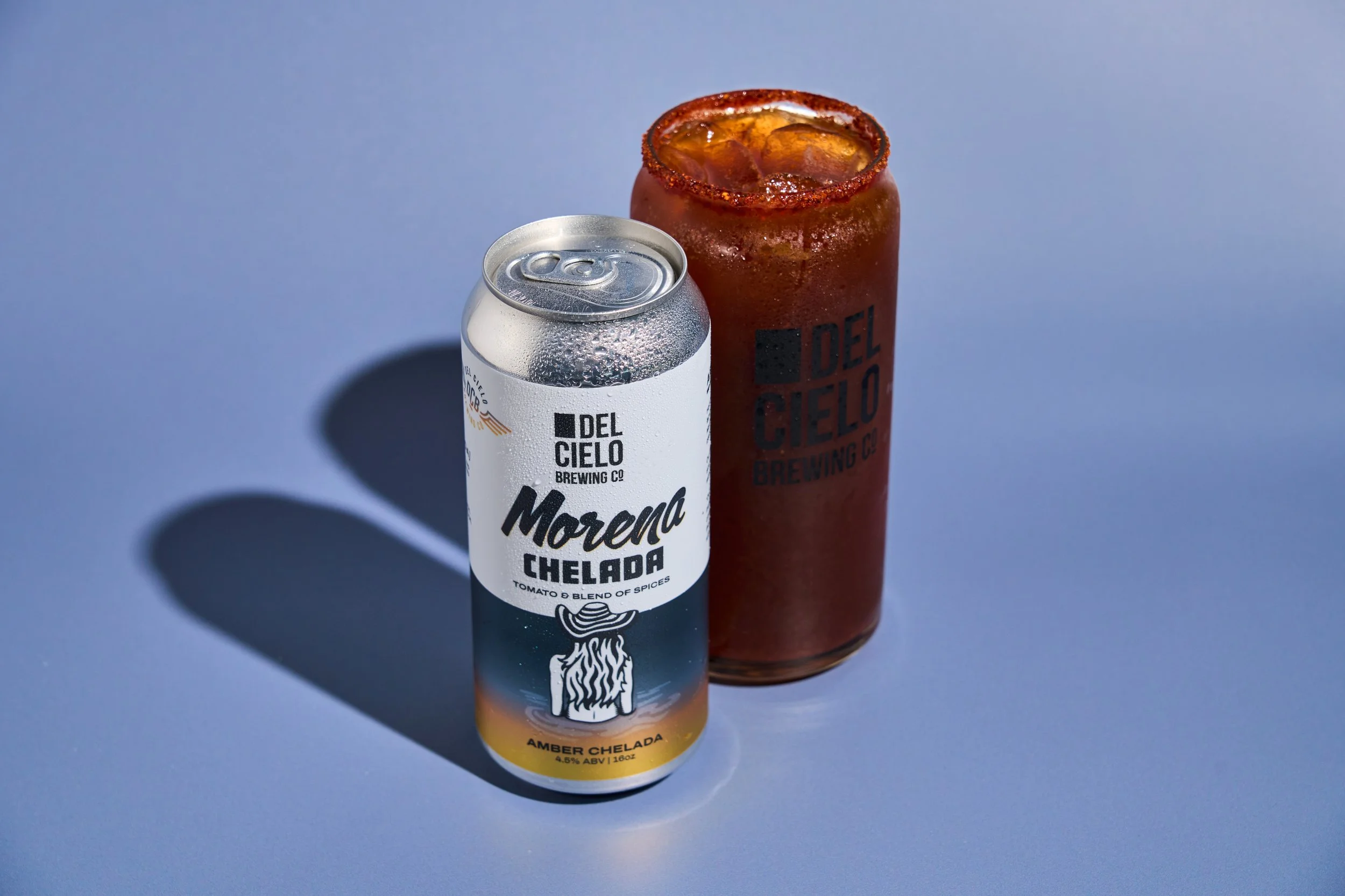 A can of Morena Chelada beer and a red glass with ice and beer, both from Del Cielo Brewing Co., on a light blue background.