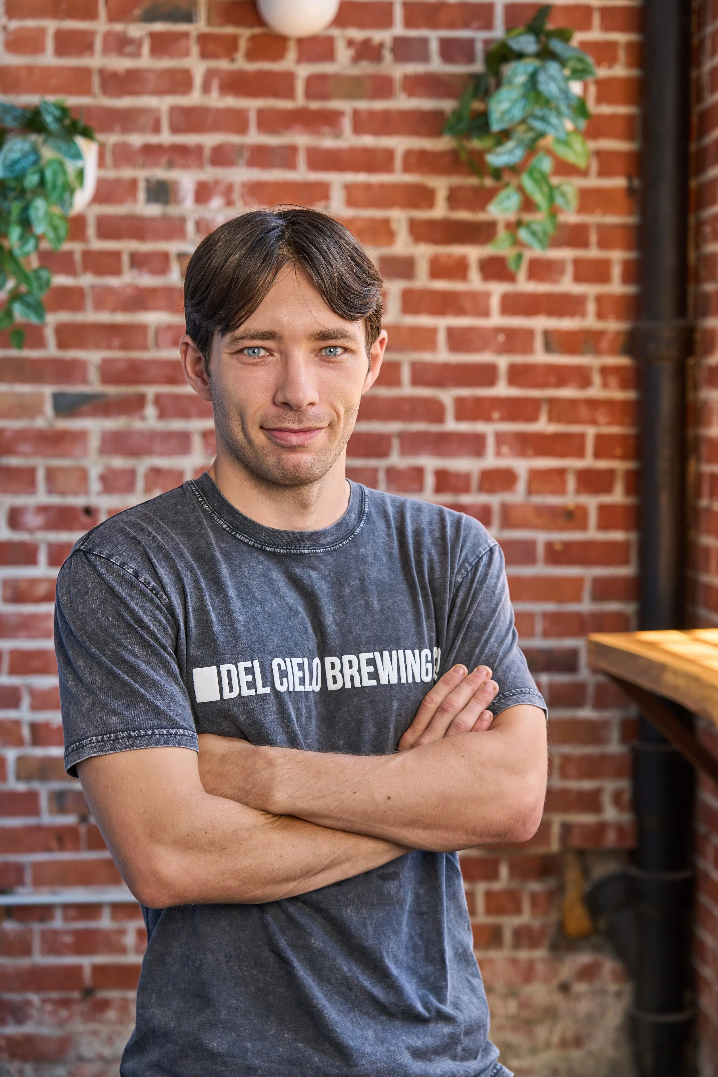 A man with dark brown hair and blue eyes crossing his arms, wearing a gray T-shirt with white text that reads 'DEL CIELO BREWING', standing in front of a red brick wall with some green plants hanging on the sides.