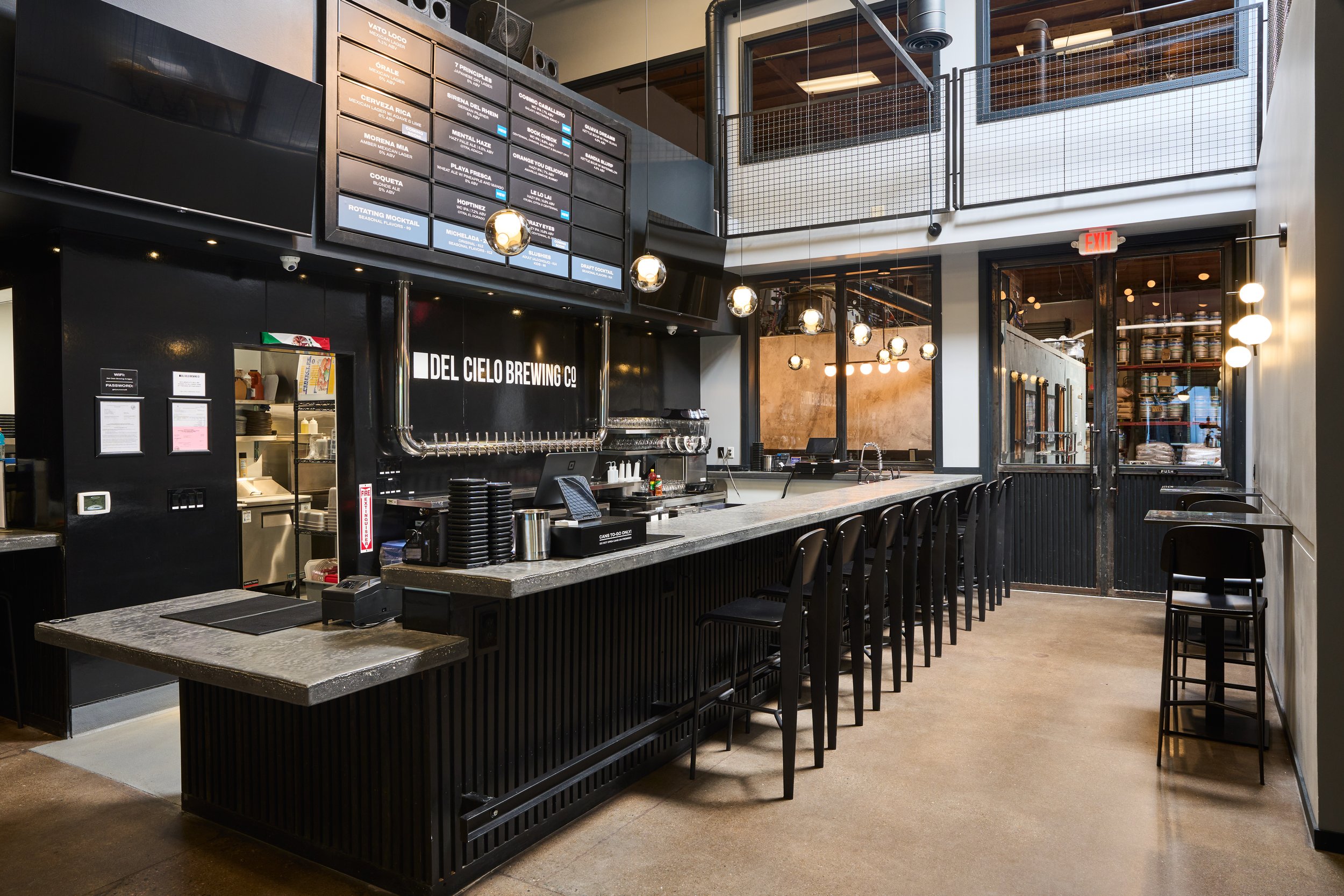 Interior of a modern brewery or bar with a black counter, high black chairs, and a large menu board overhead listing various drinks. There is a brewing setup behind the counter, and the decor includes hanging spherical lights and a loft area with metal railings. The space has a contemporary industrial design.