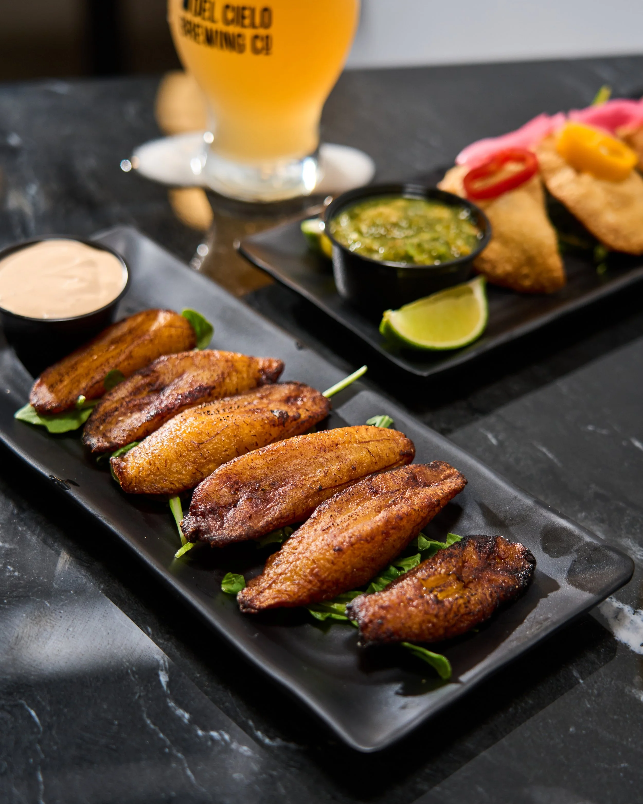 Close-up of grilled chicken wings with seasoning, served on a black rectangular plate with greens underneath, accompanied by lime wedges, in front of a black stone surface.
