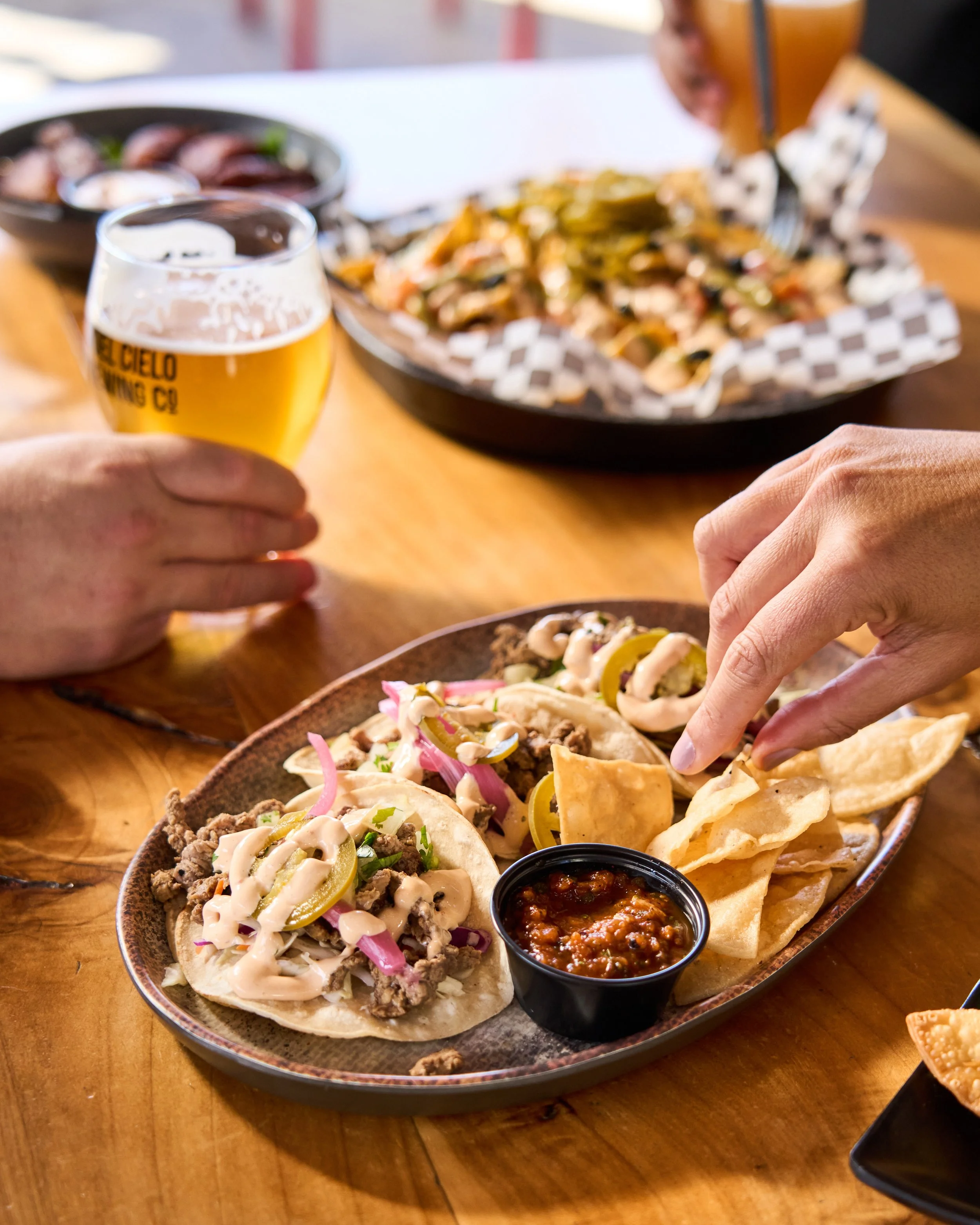 A tray of Mexican tacos with various toppings including meat, onions, cilantro, and sliced jalapenos, accompanied by a small cup of salsa on a wooden table.