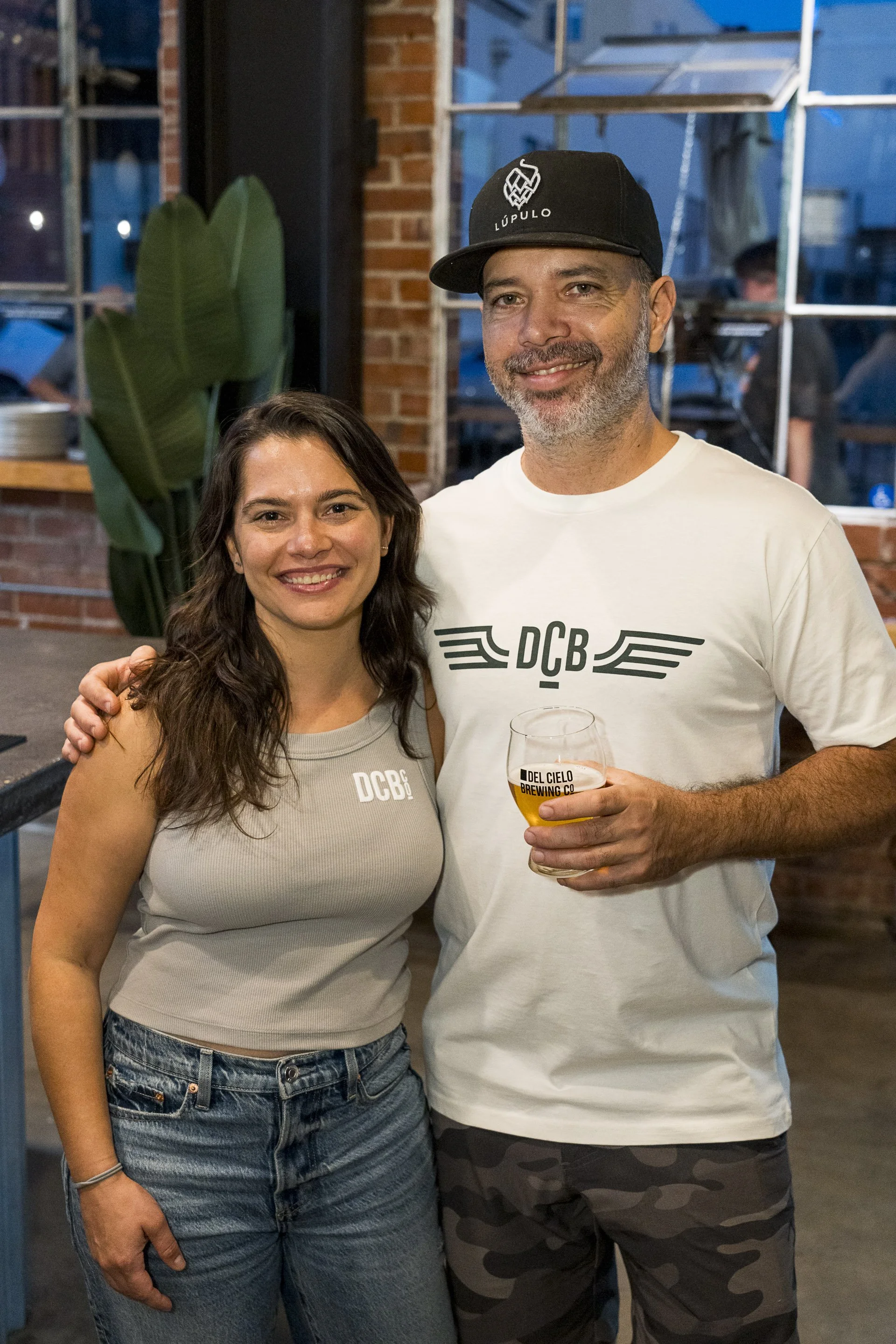 A woman and a man smiling and posing together at a bar or brewery. The woman is wearing a light gray tank top and denim jeans, the man is wearing a white t-shirt with a logo and camo shorts, and holding a glass of beer. The background shows brick wal