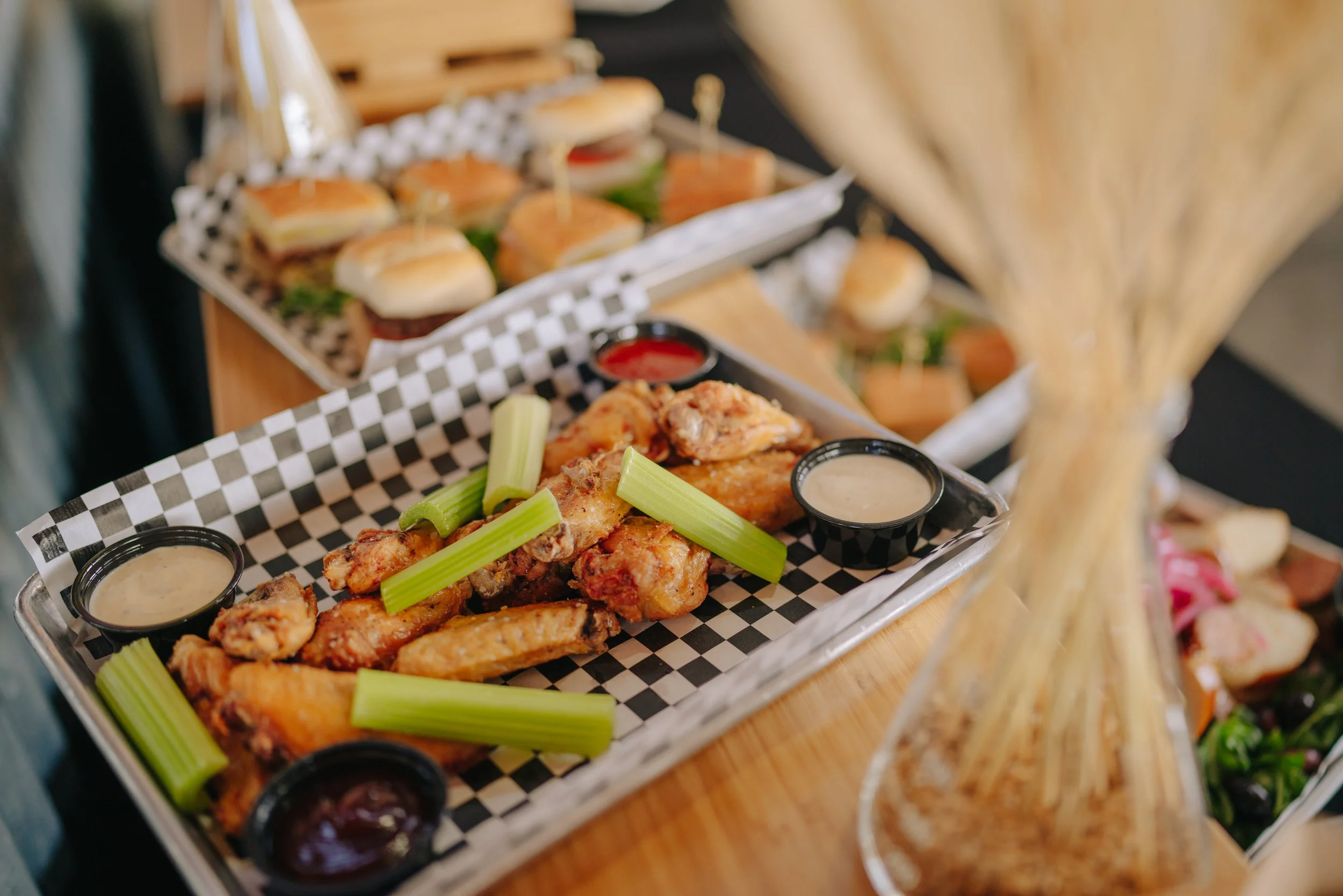 Basket of fried chicken wings with celery sticks and dipping sauces, with a tray of mini sandwiches in the background.