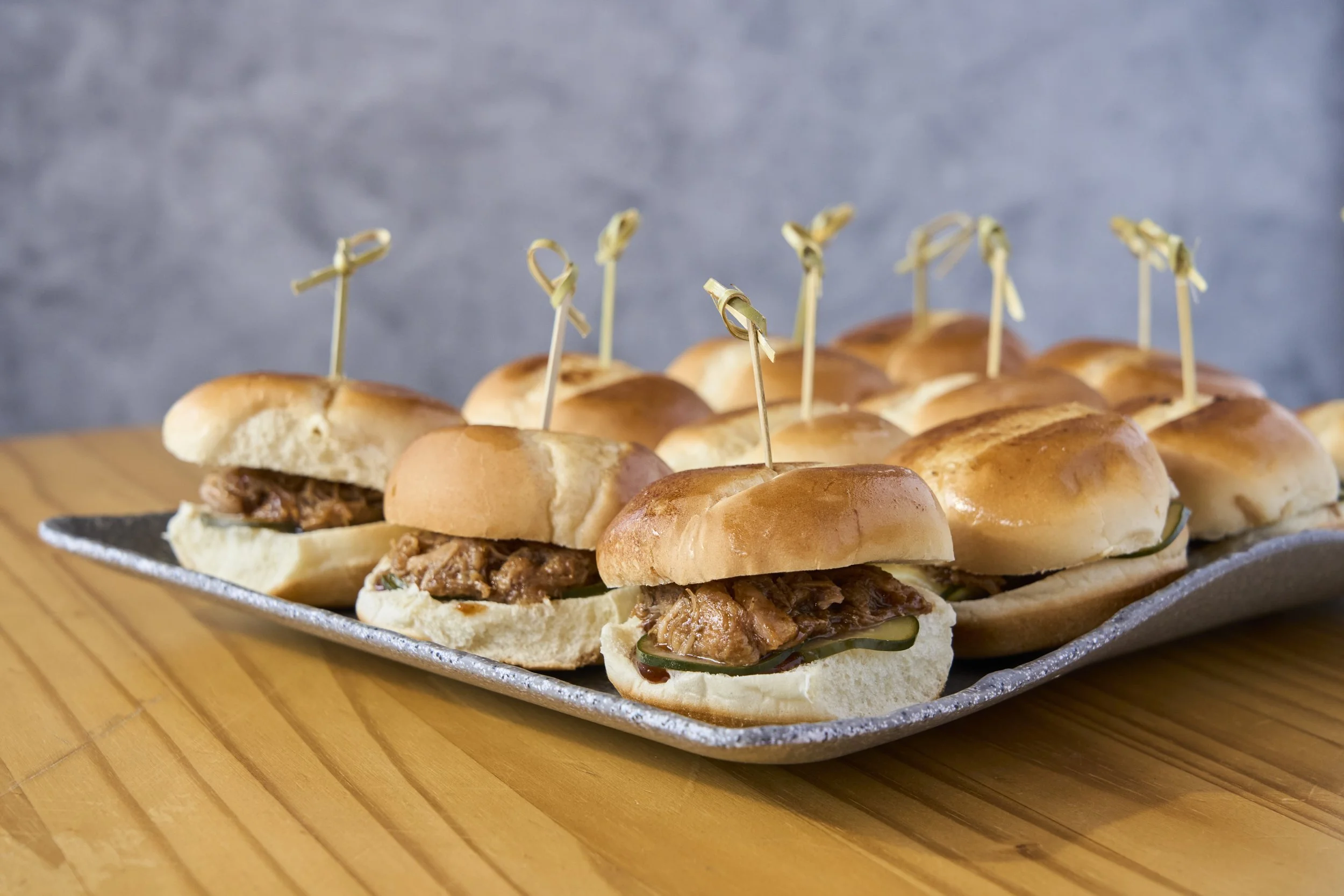 A tray of small sandwiches with pulled pork, topped with small skewers, on a wooden surface.