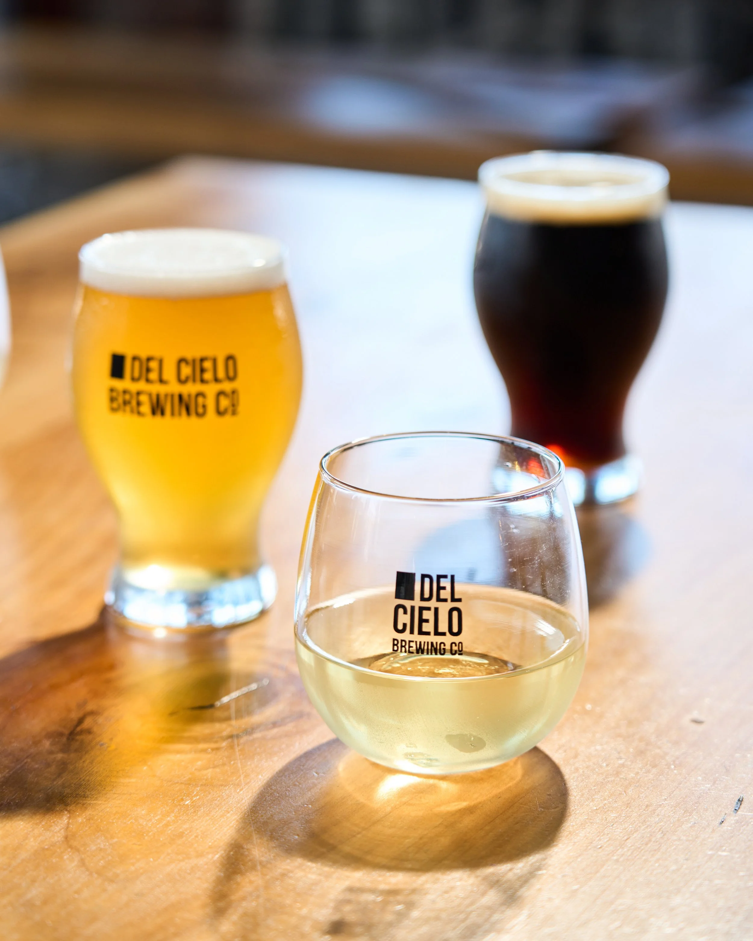 Two pints of beer on a wooden table, with the logos ’Del Cielo Brewing Co’ and a glass of white wine in the foreground.