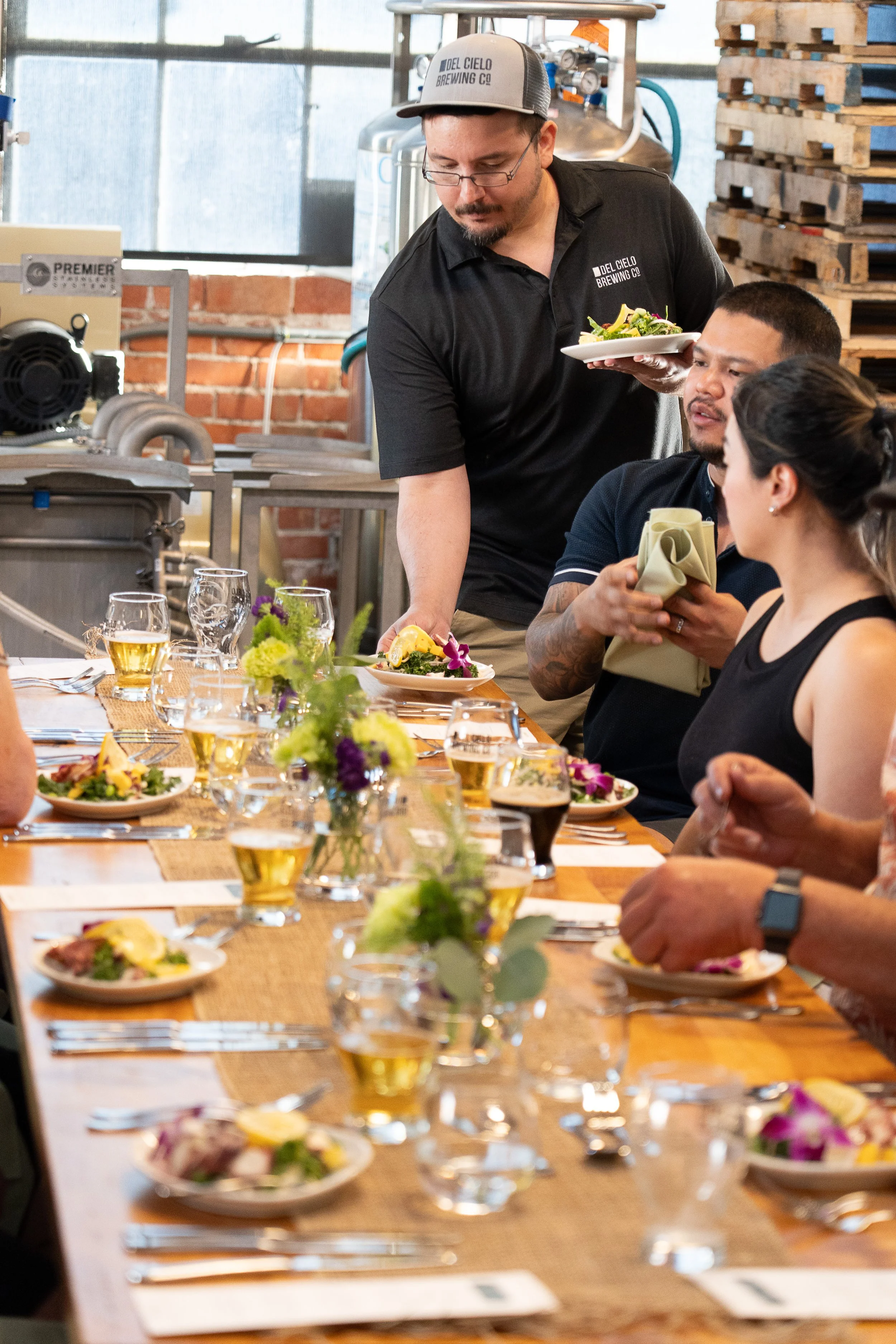 A group of people sitting at a long restaurant table with plates of food, drinks, and floral centerpieces, while a waiter serves a salad in a serving dish.