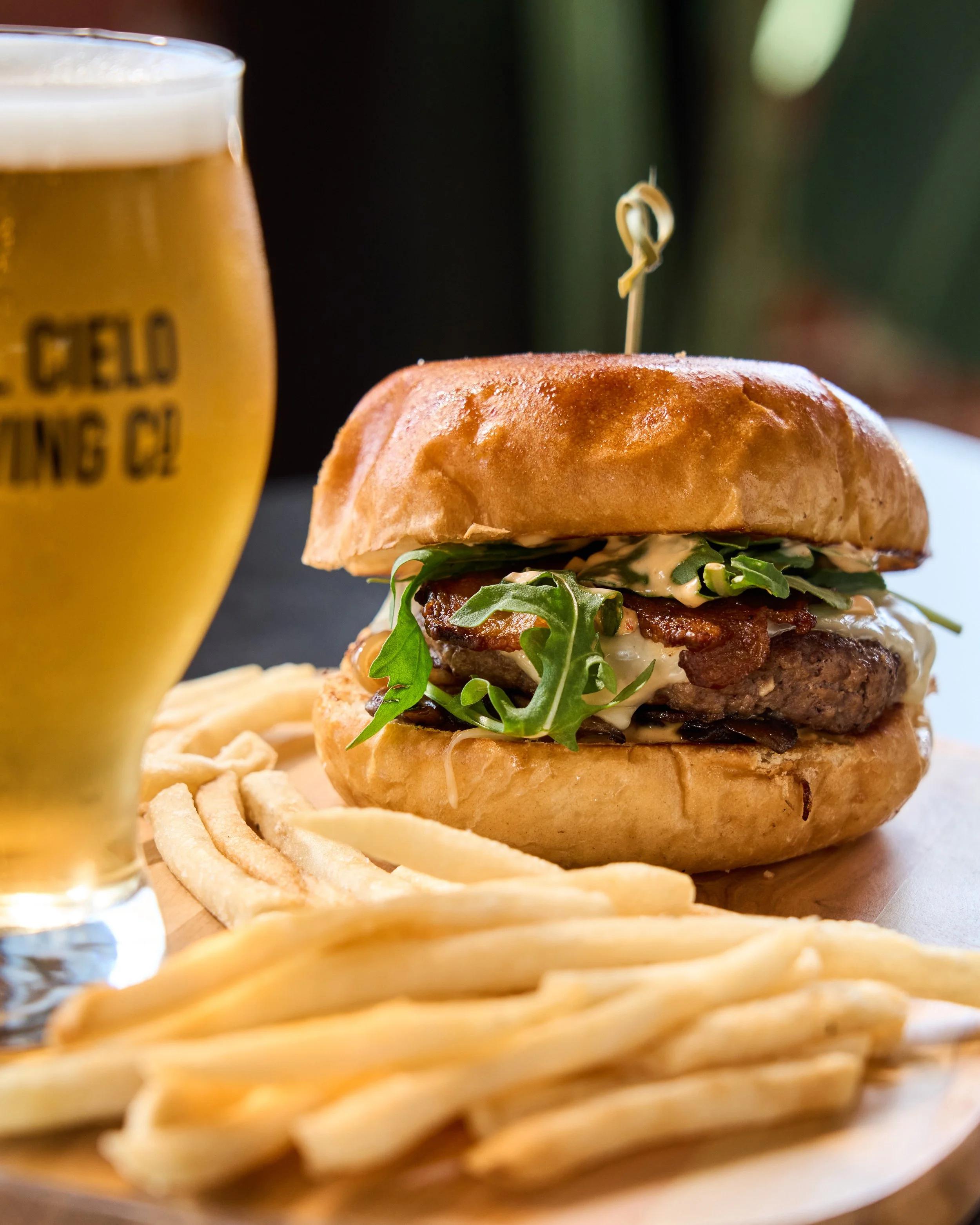 A close-up of a cheeseburger with lettuce, bacon, and sauce, sitting on a bun with a skewer on top, accompanied by shoestring fries and a glass of beer in the background.