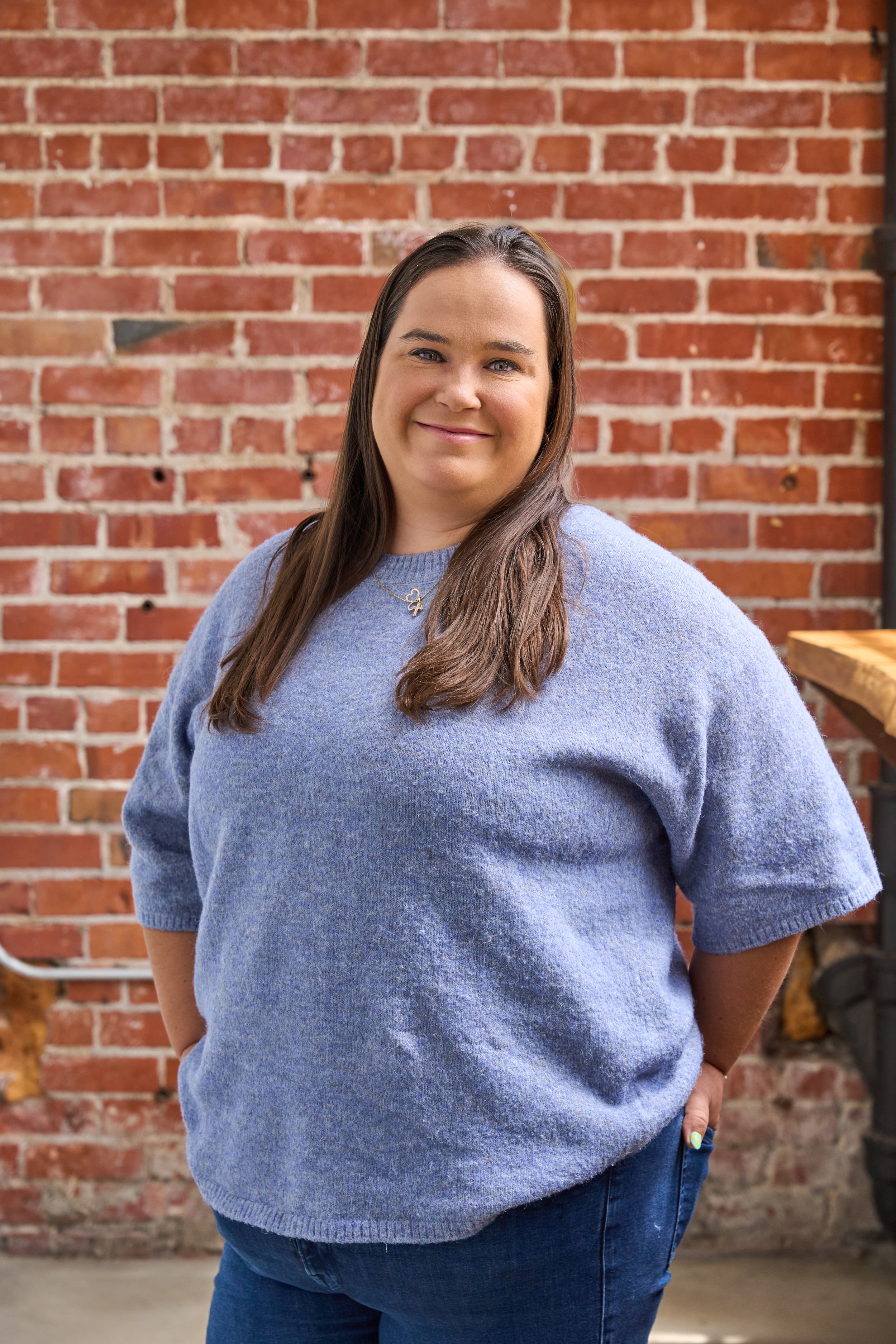 A woman with long brown hair smiling, wearing a blue sweater and jeans, standing in front of a brick wall decorated with a large lit arrow sign and green ivy.