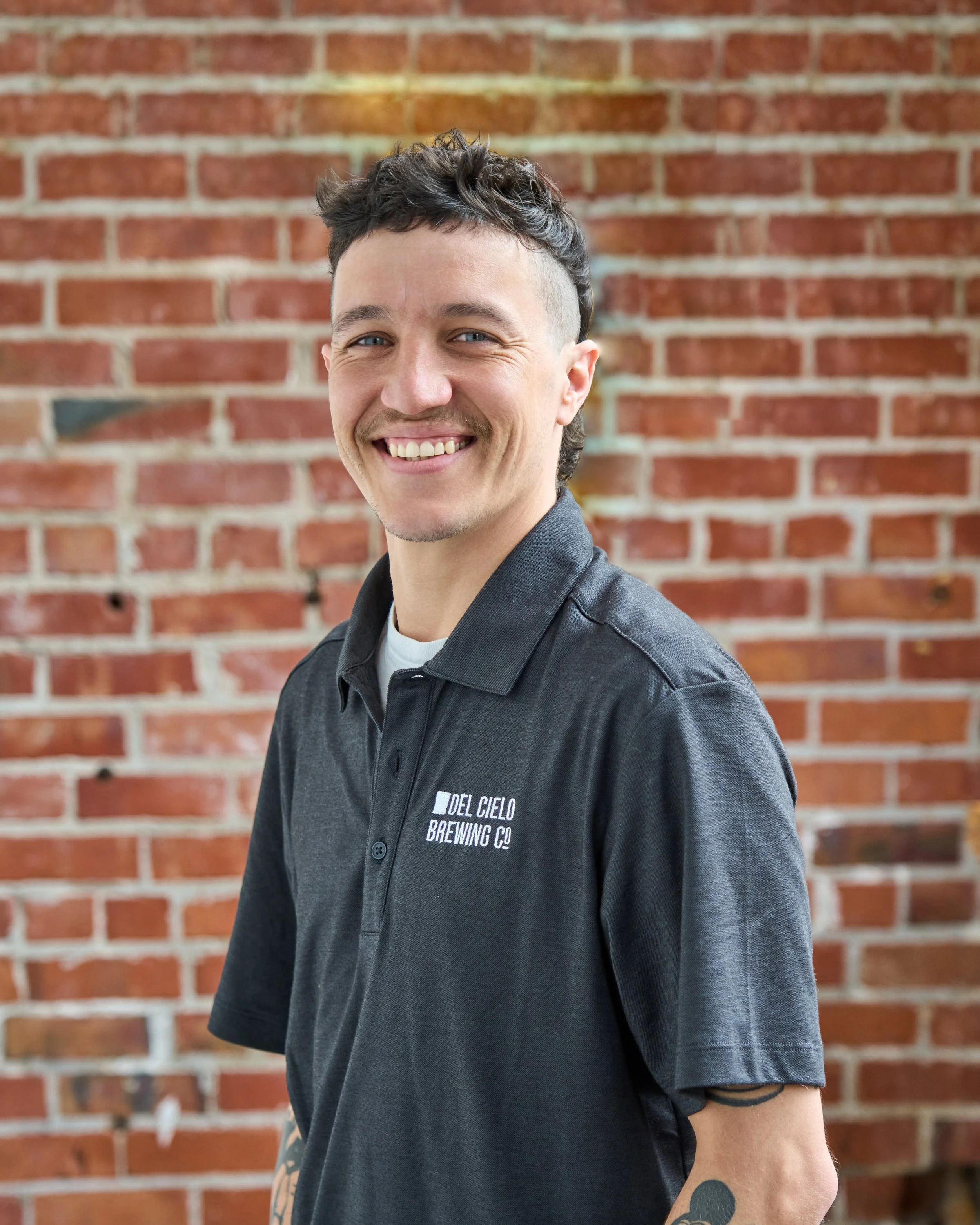 A smiling young man with tattoos on his left arm stands in front of a brick wall with a large, yellow marquee number 7 outlined with lights, along with some green plants hanging on the wall. He wears a black polo shirt with a logo and light blue jeans.