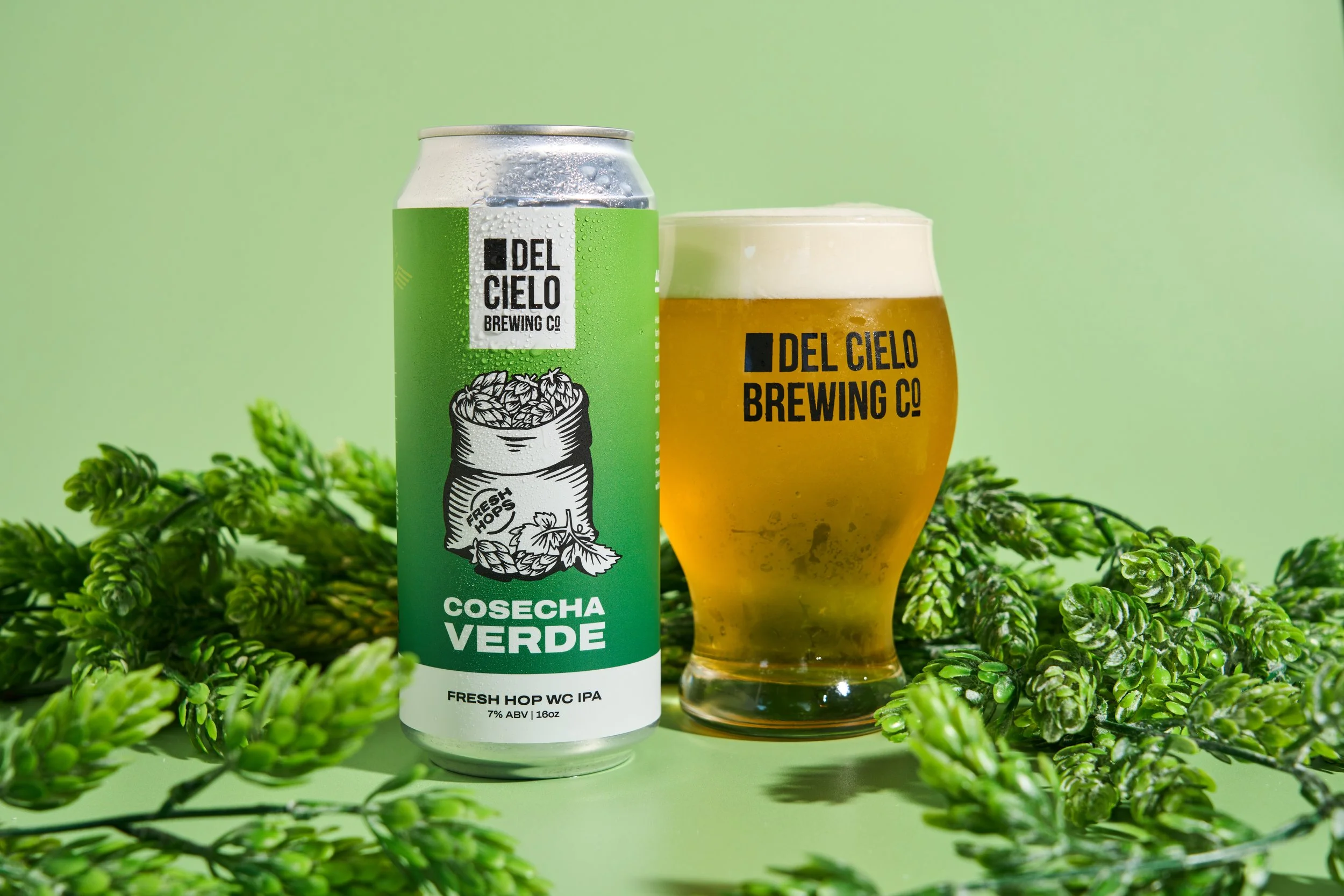 A can of Del Cielo Cosecha Verde Fresh Hop WC IPA beer next to a glass filled with beer, surrounded by green hops on a light green background.