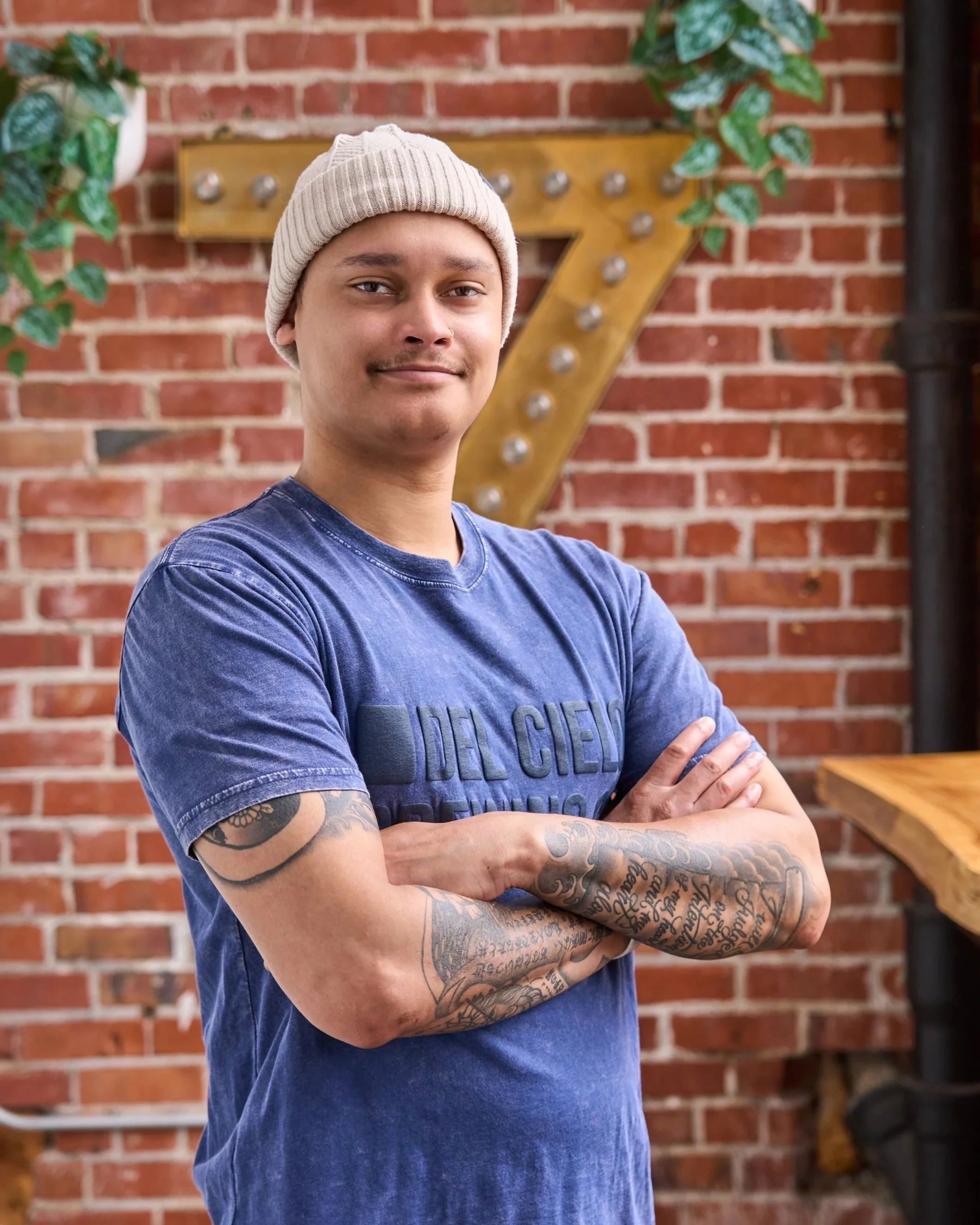 A young man with tattoos on his arms wearing a beige beanie and a blue T-shirt standing outdoors against a brick wall with a large vintage-style letter '7' sign and some greenery.