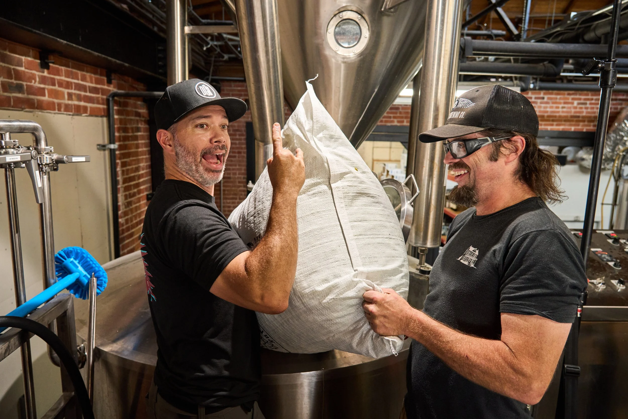 Two men are in a brewery, one is holding a large bag of ingredients while the other is smiling and pointing upward. They are standing near brewing equipment and brick walls.