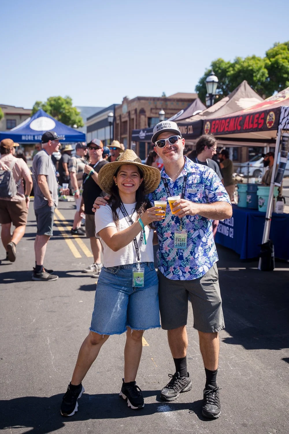 Two people, a woman and a man, smiling and holding cups of beer at an outdoor event with tents and people in the background.