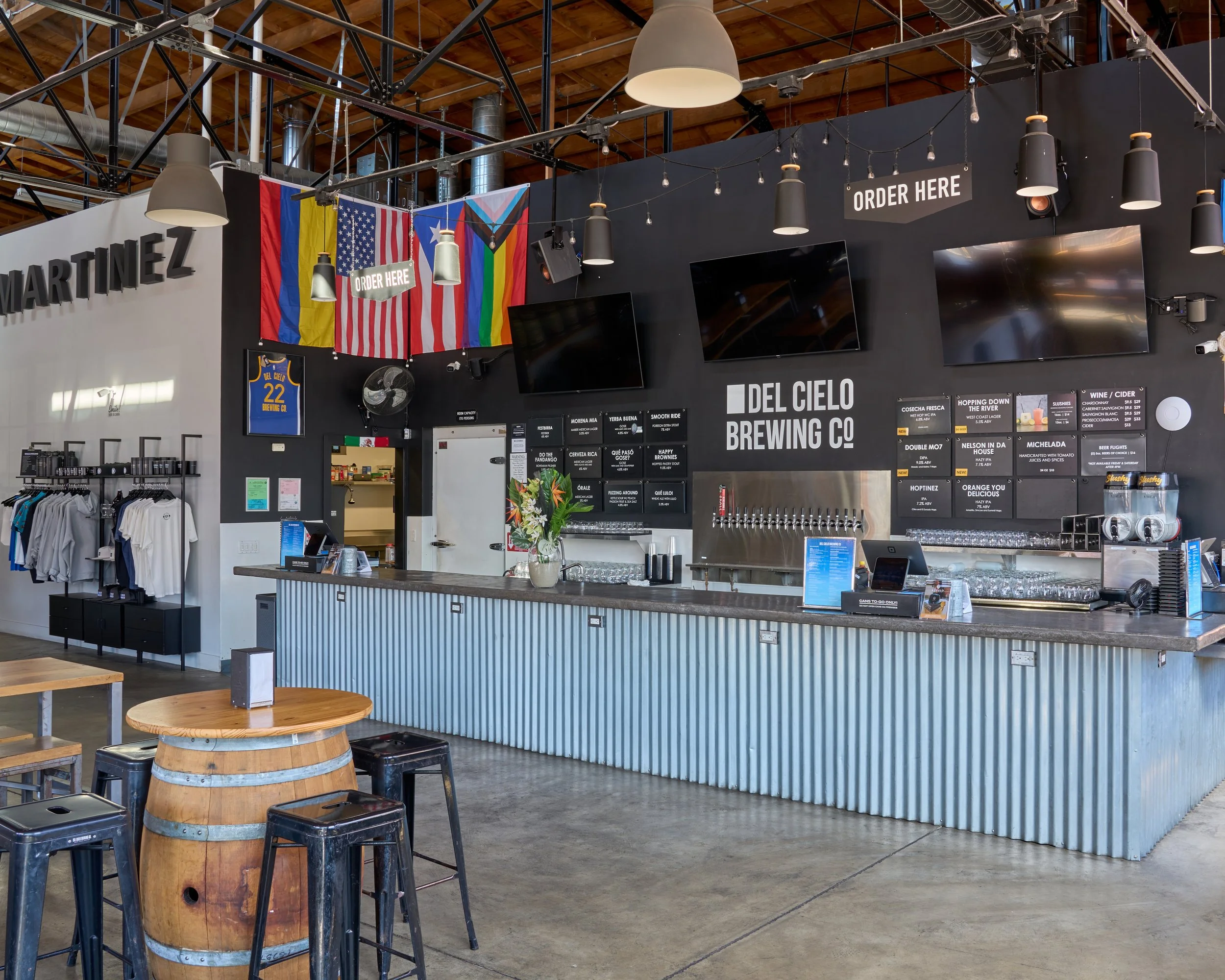 Inside a modern brewery or bar with a black wall, multiple flat-screen TVs, and signs indicating where to order; colorful flags and banners hang from the ceiling; a counter with a vase of flowers and glassware; wooden tables and barrel-shaped stools 