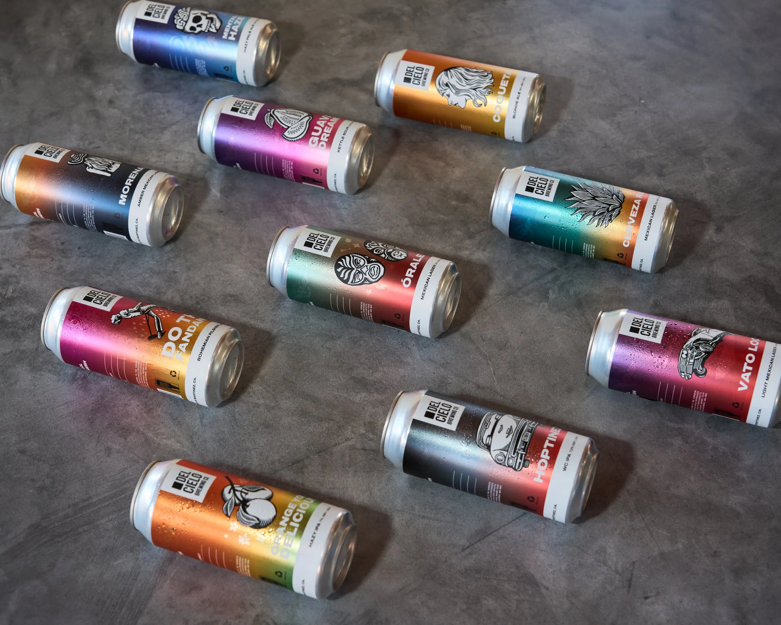 A collection of colorful cans of Del Cielo beer arranged on a gray surface.