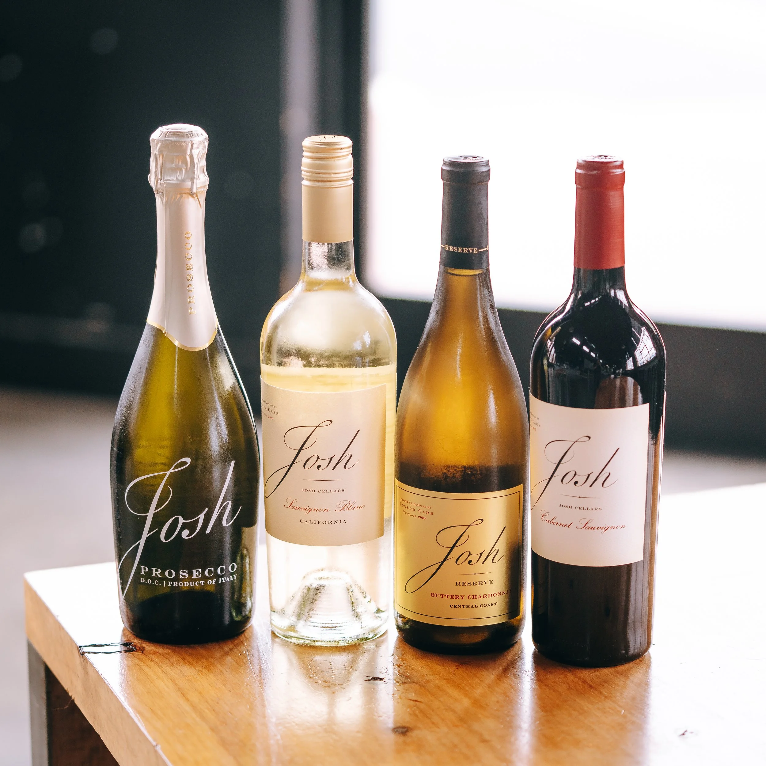 Four bottles of wine on a wooden surface with a blurred background, featuring close-up views of their necks and labels.