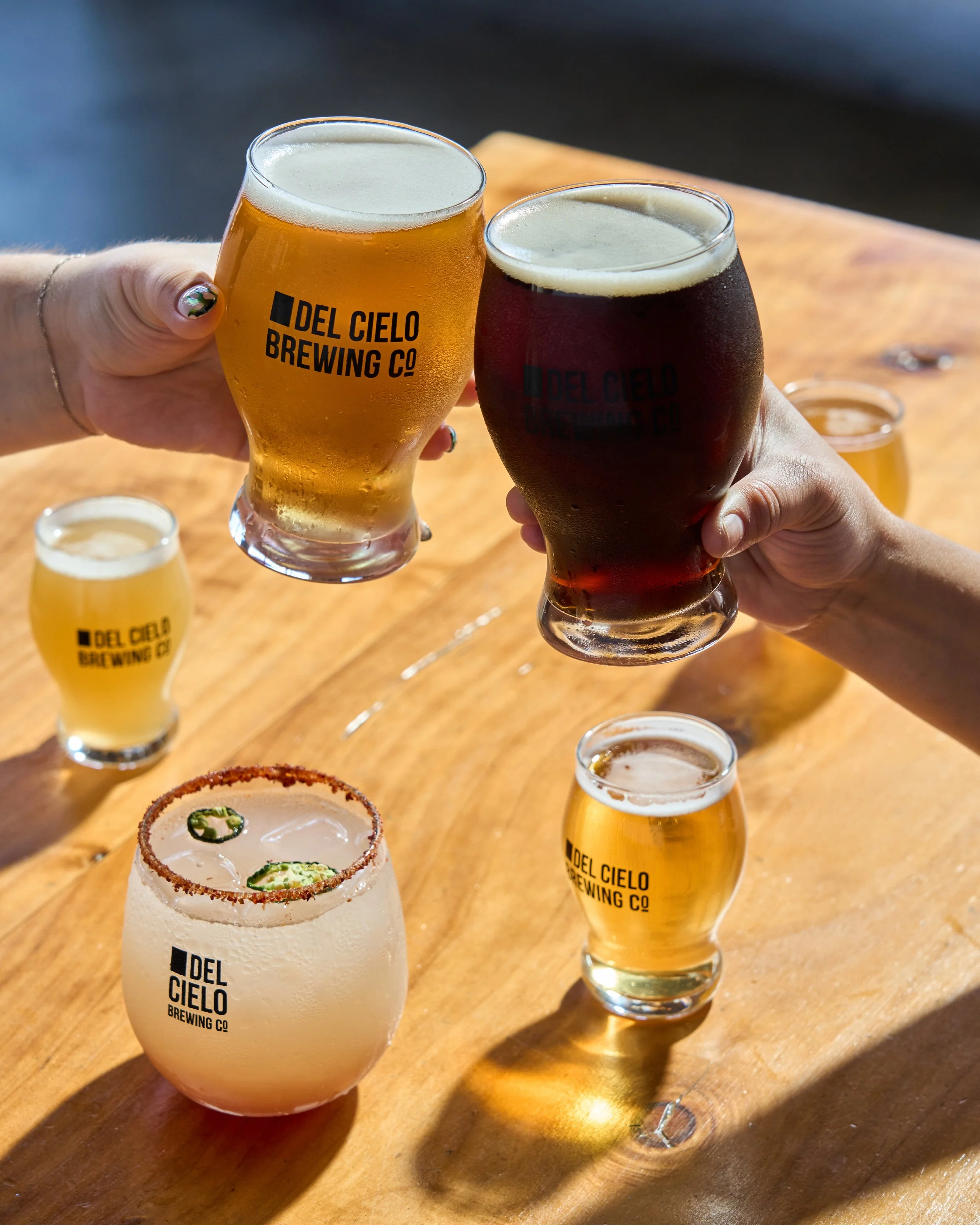 Two people clinking glasses of beer, one light and one dark, at a wooden table with additional beer glasses and a margarita cocktail in the foreground. The glasses have the logo 'DEL CIELO BREWING CO'.