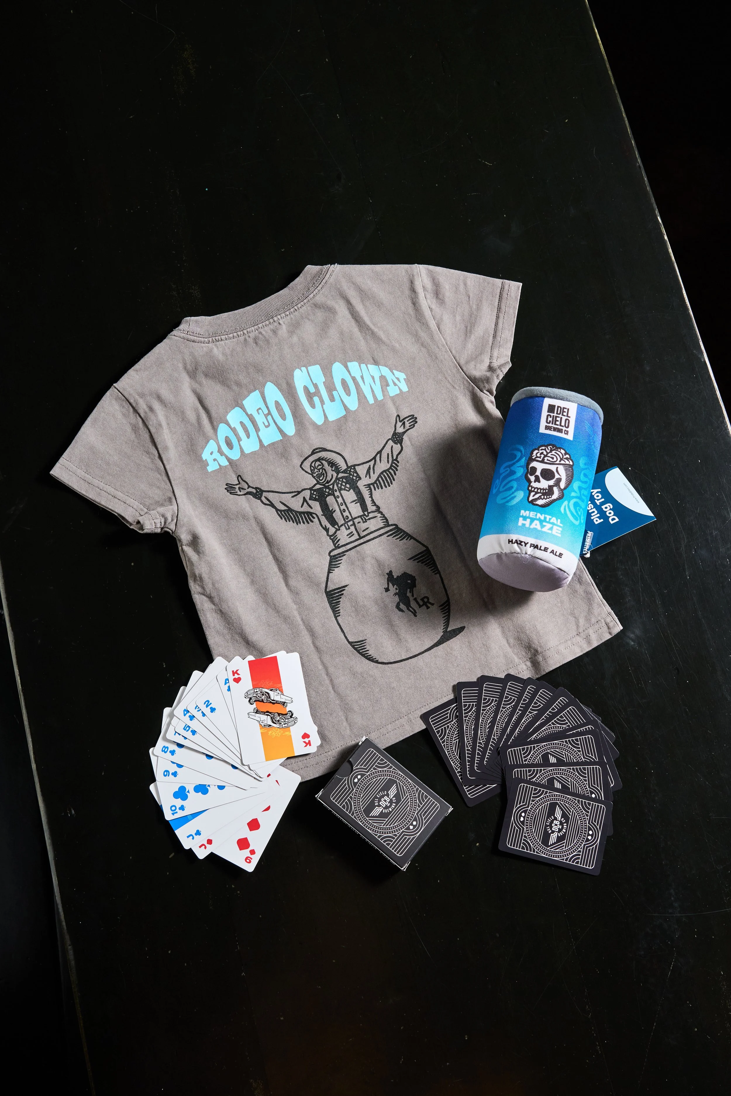 A table with a gray T-shirt featuring a graphic design and text, a can of Del Cielo beer labeled 'Mental Haze Hazy Pale Ale,' a deck of playing cards, and multiple black-backed cards.