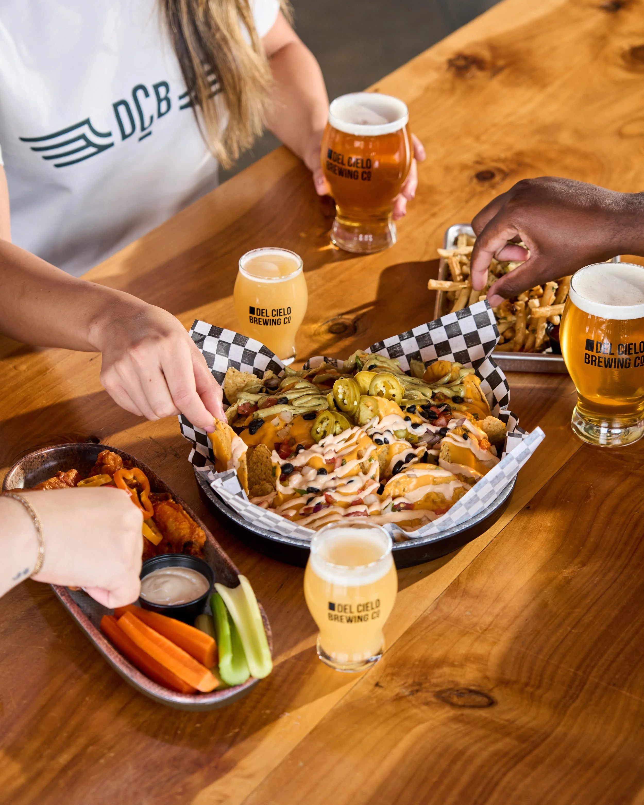 People sharing a nachos and drinks at a wooden table in a restaurant or bar. There are two glasses of beer with 'Del Cielo Brewing Co.' written on them, and a tray of vegetables and dipping sauces.