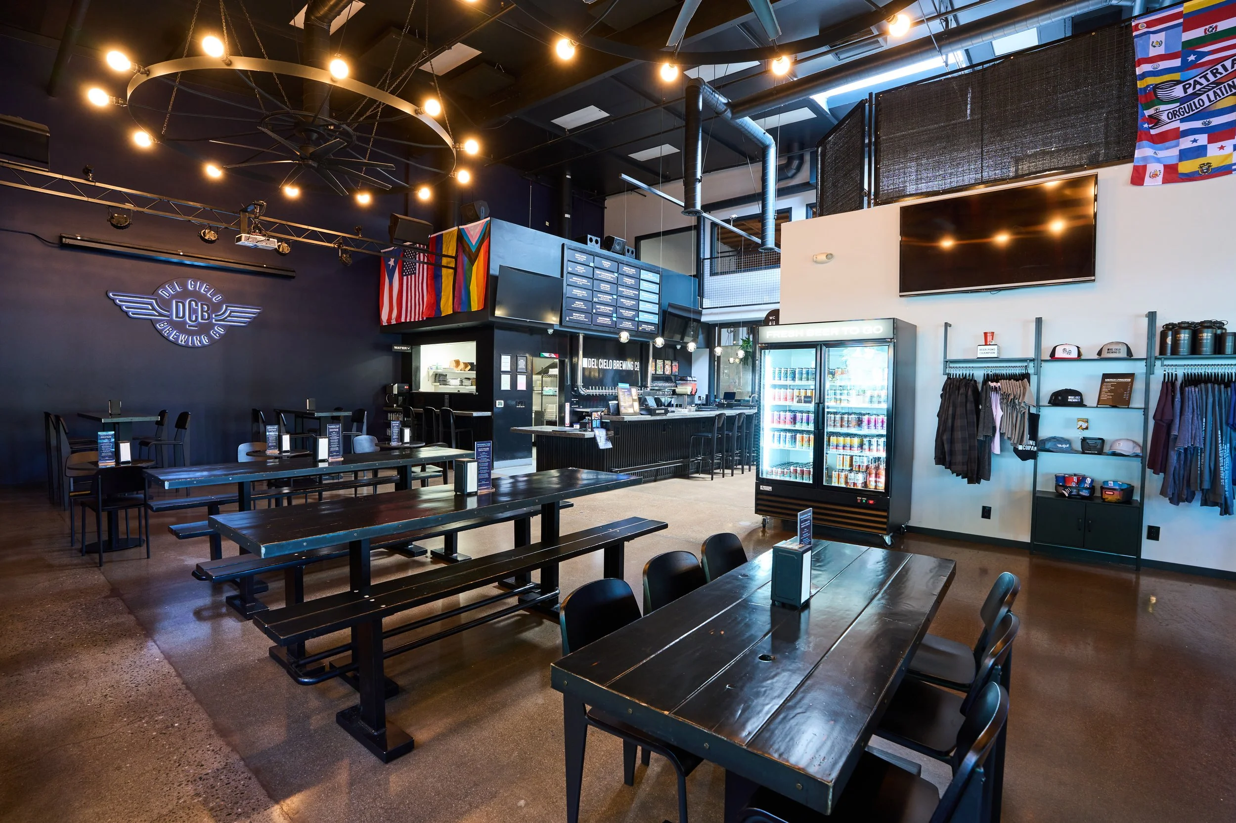 Interior of a modern bar or restaurant with black tables and chairs, a dark blue wall with a neon sign, a large flat-screen TV, and flags, including a rainbow pride flag and a Puerto Rican flag. There is a refrigerated display case with drinks and various merchandise.