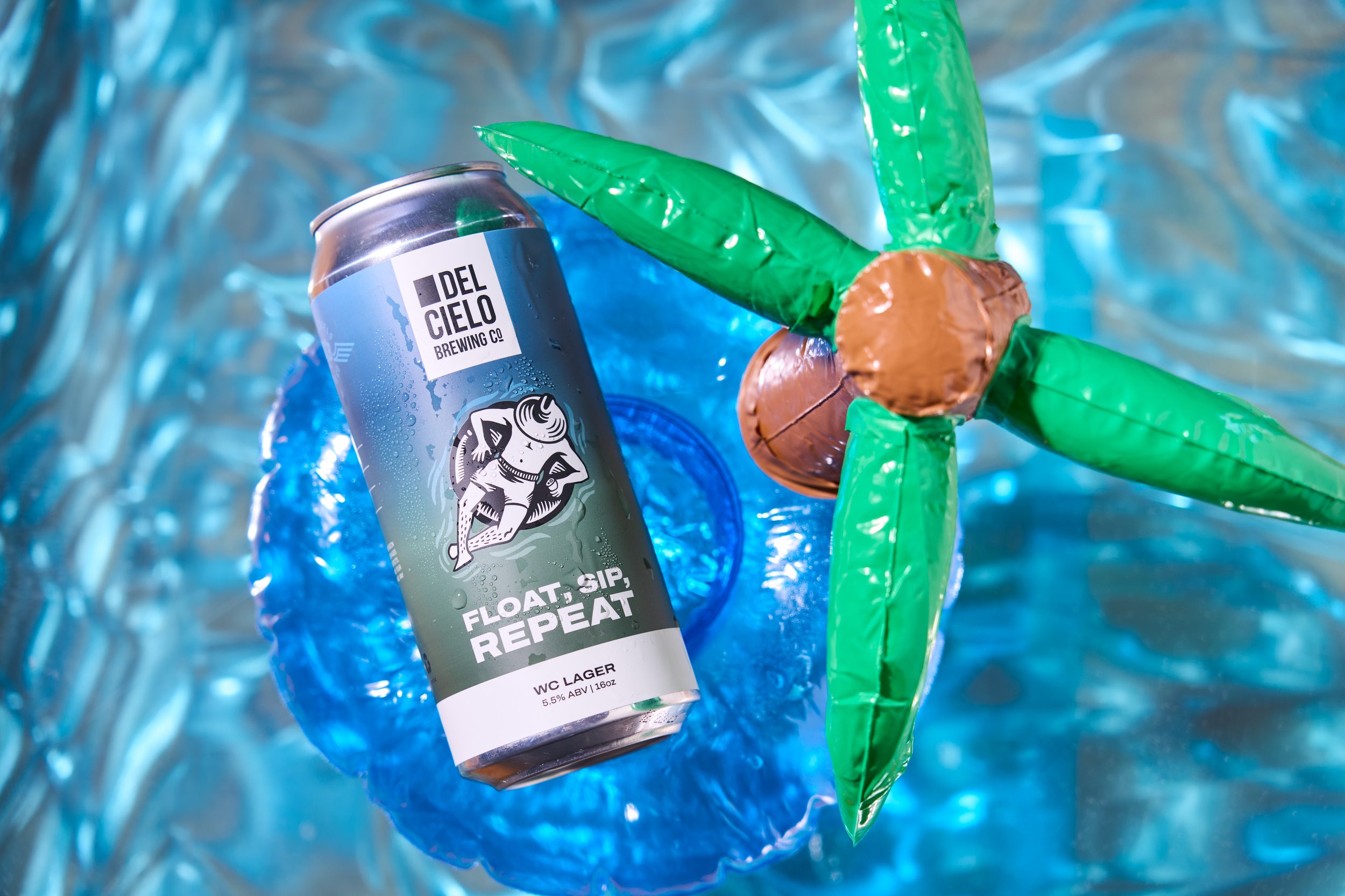 Can of Del Cielo Brewing Co. beer with a label featuring a black and white illustration of a man on a floatie and the text 'Float, Sip, Repeat' surrounded by green inflatable palm trees and brown coconuts on a blue inflatable background.