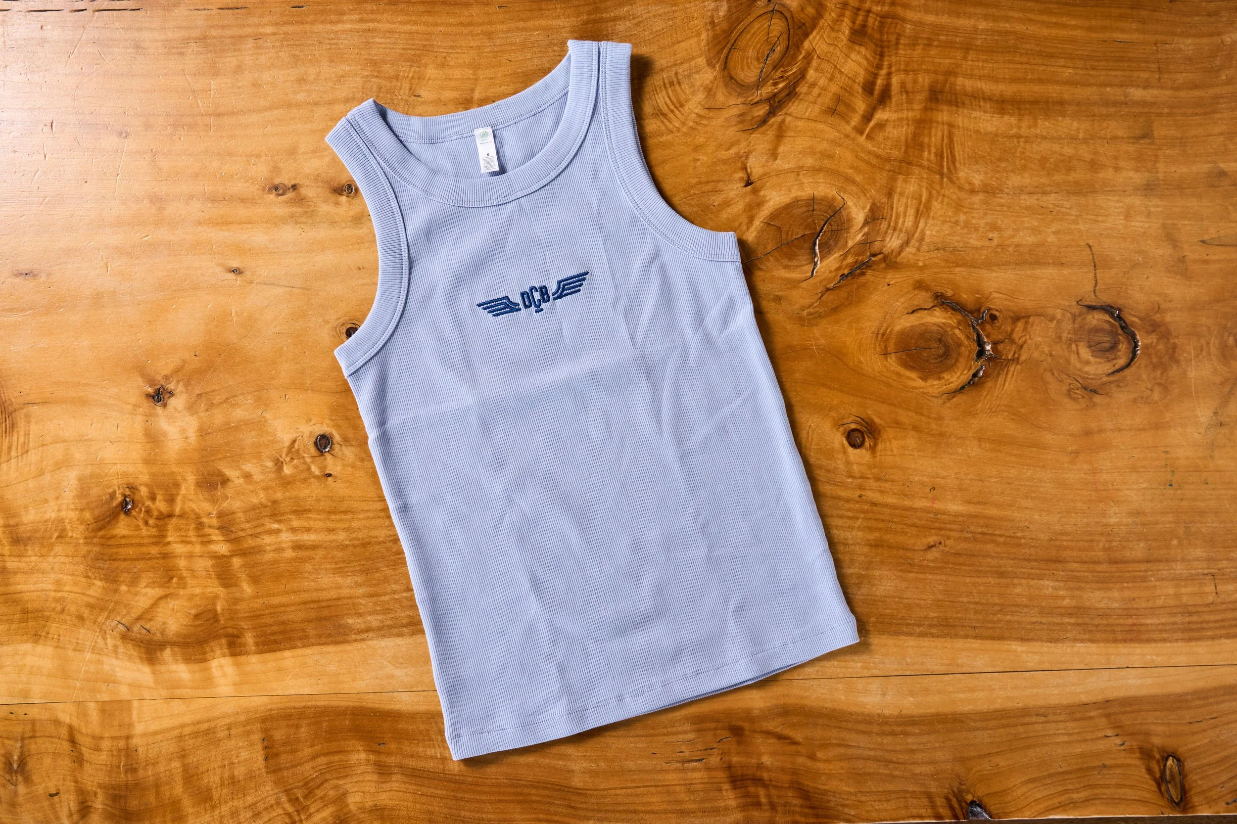 A light blue sleeveless tank top with embroidered dark blue letters "DCB" and wings is placed on a wooden surface with visible knots and grain.