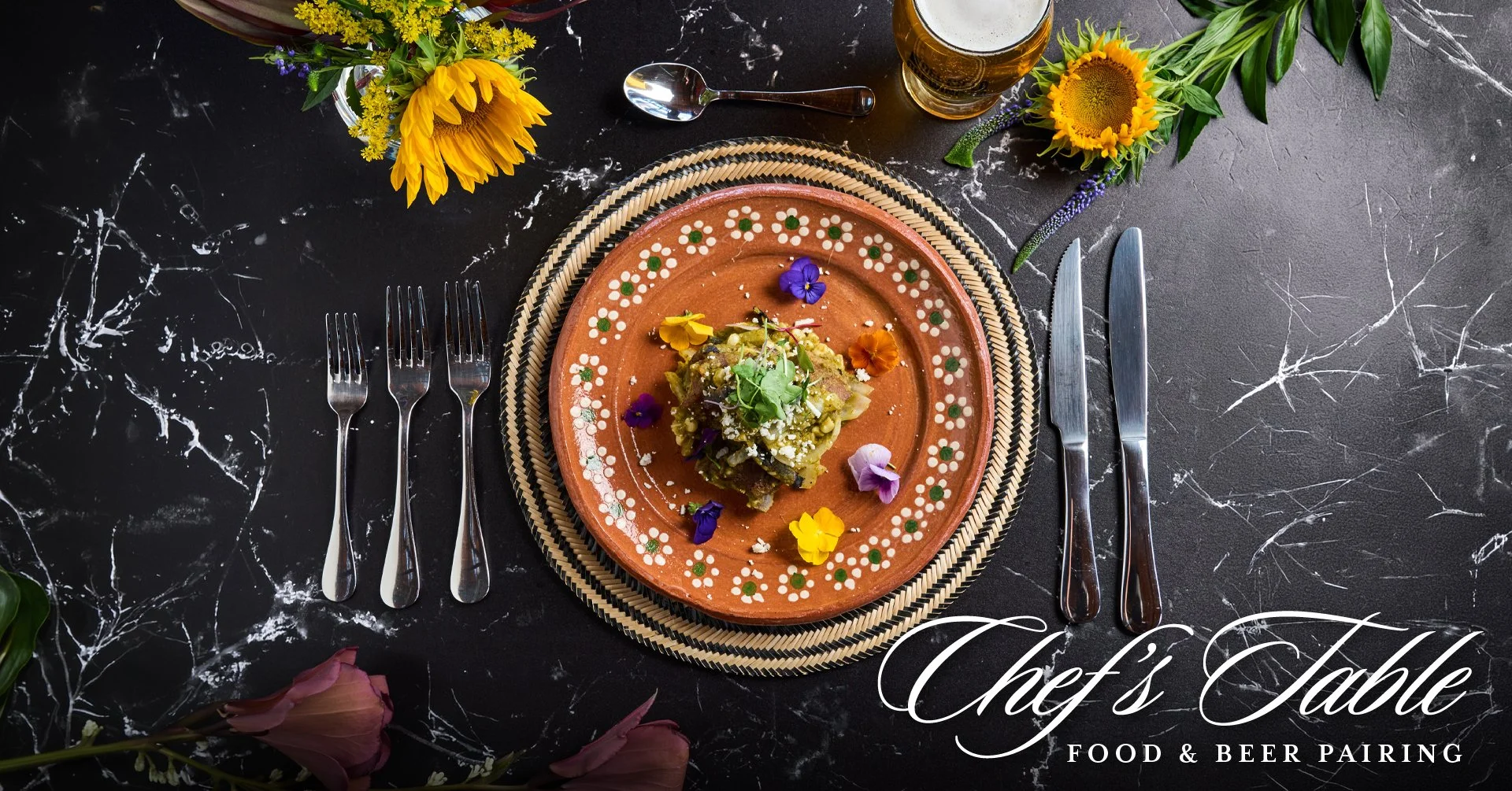 A colorful table setting featuring a terracotta plate with food garnished with edible flowers, set on a woven placemat, flanked by silverware, a spoon, and a knife. Bright yellow sunflowers and other flowers decorate the table, which has a dark marble surface. There's a glass of beer and additional flowers in the background.