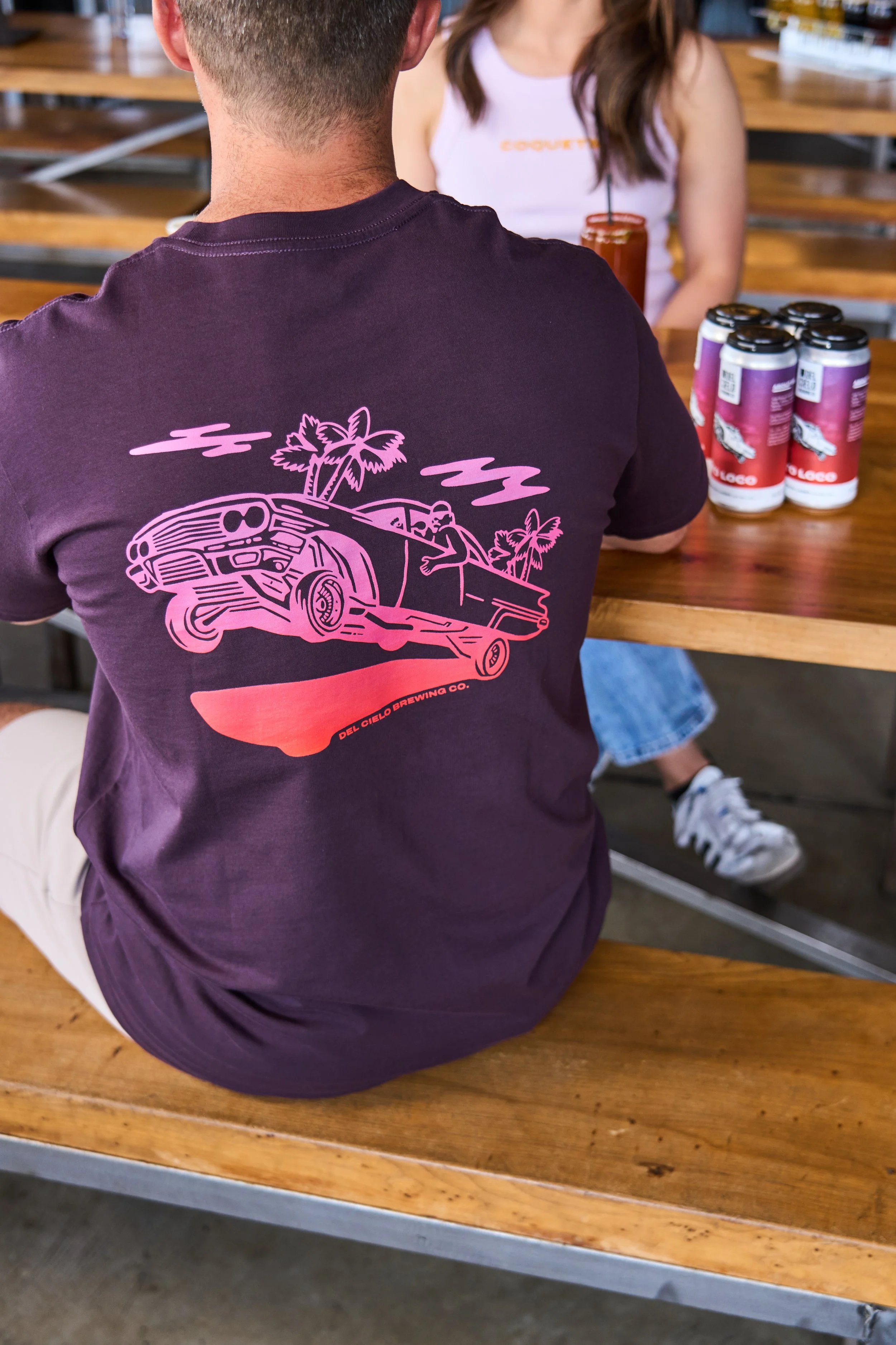 A person wearing a purple t-shirt with a graphic of a low-rider car, palm trees, and clouds, sitting at a wooden table with cans of drinks and a woman with long hair in a white tank top in the background.