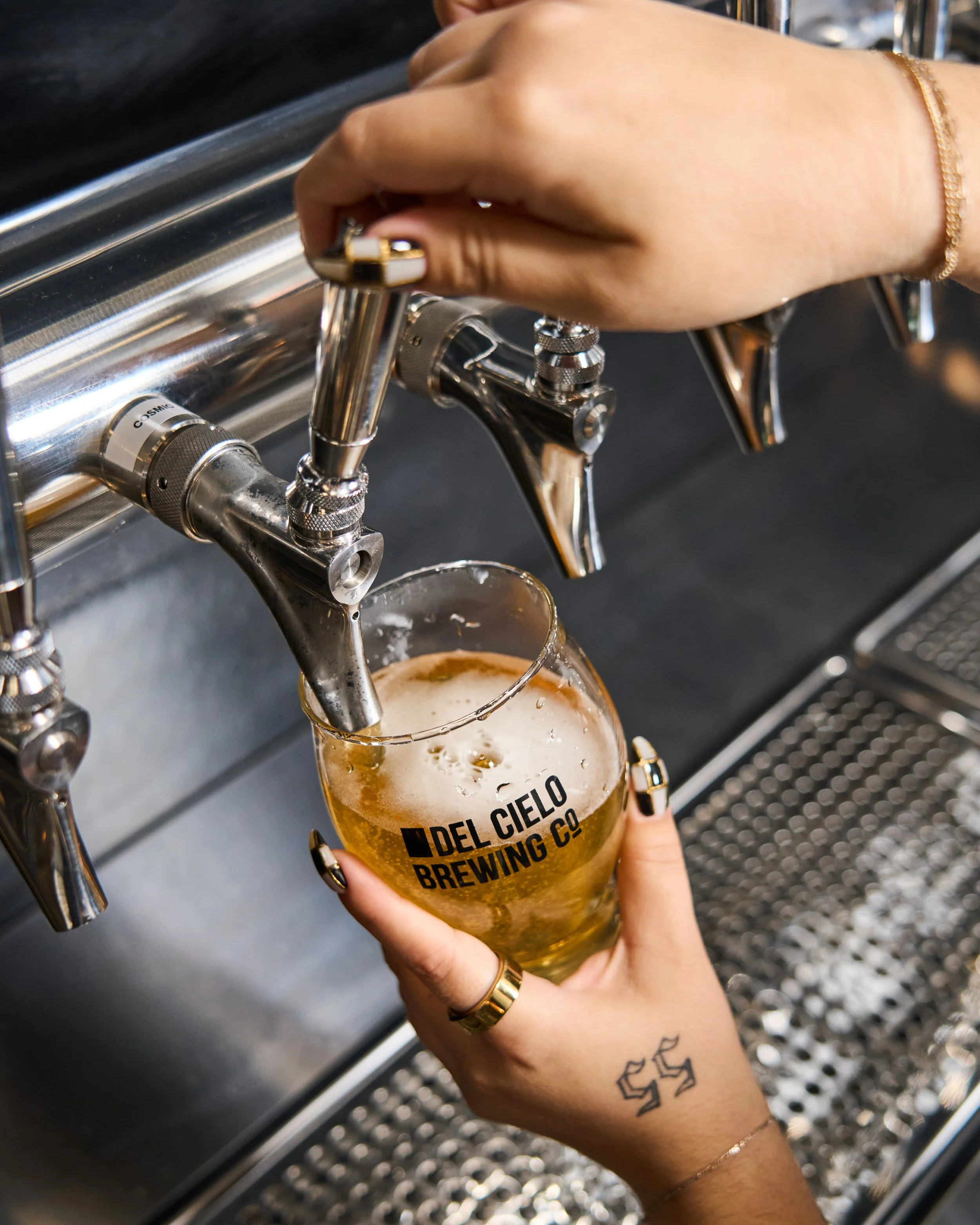 A person with tattoos on their wrist filling a glass of beer from a tap at Del Cielo Brewing Co.