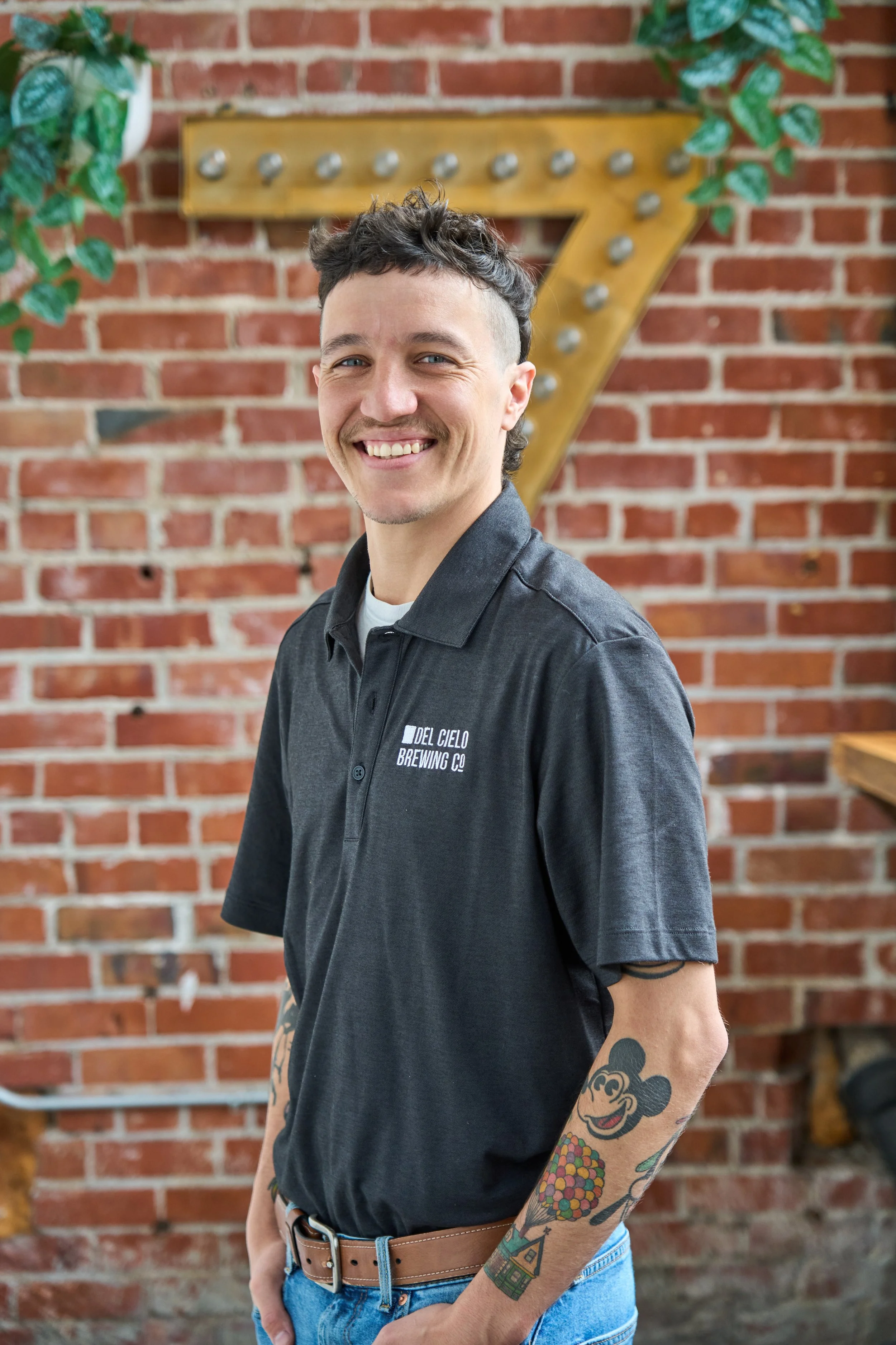 A smiling young man with tattoos on his left arm stands in front of a brick wall with a large, yellow marquee number 7 outlined with lights, along with some green plants hanging on the wall. He wears a black polo shirt with a logo and light blue jeans.
