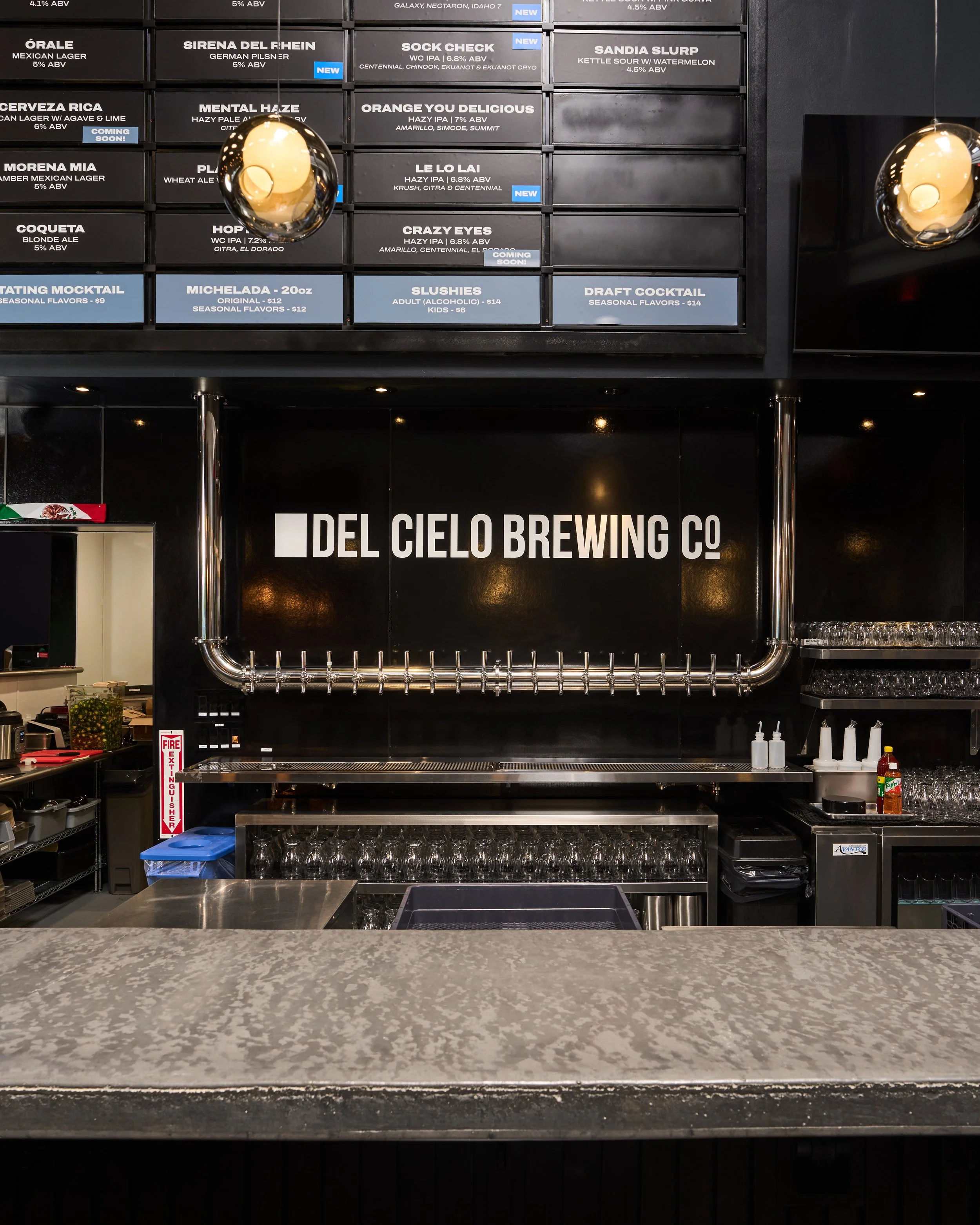 Bar counter with empty glasses, bottles, and condiment bottles, in front of a black wall with a sign reading 'Del Cielo Brewing Co.' and a menu board with various beers and prices.