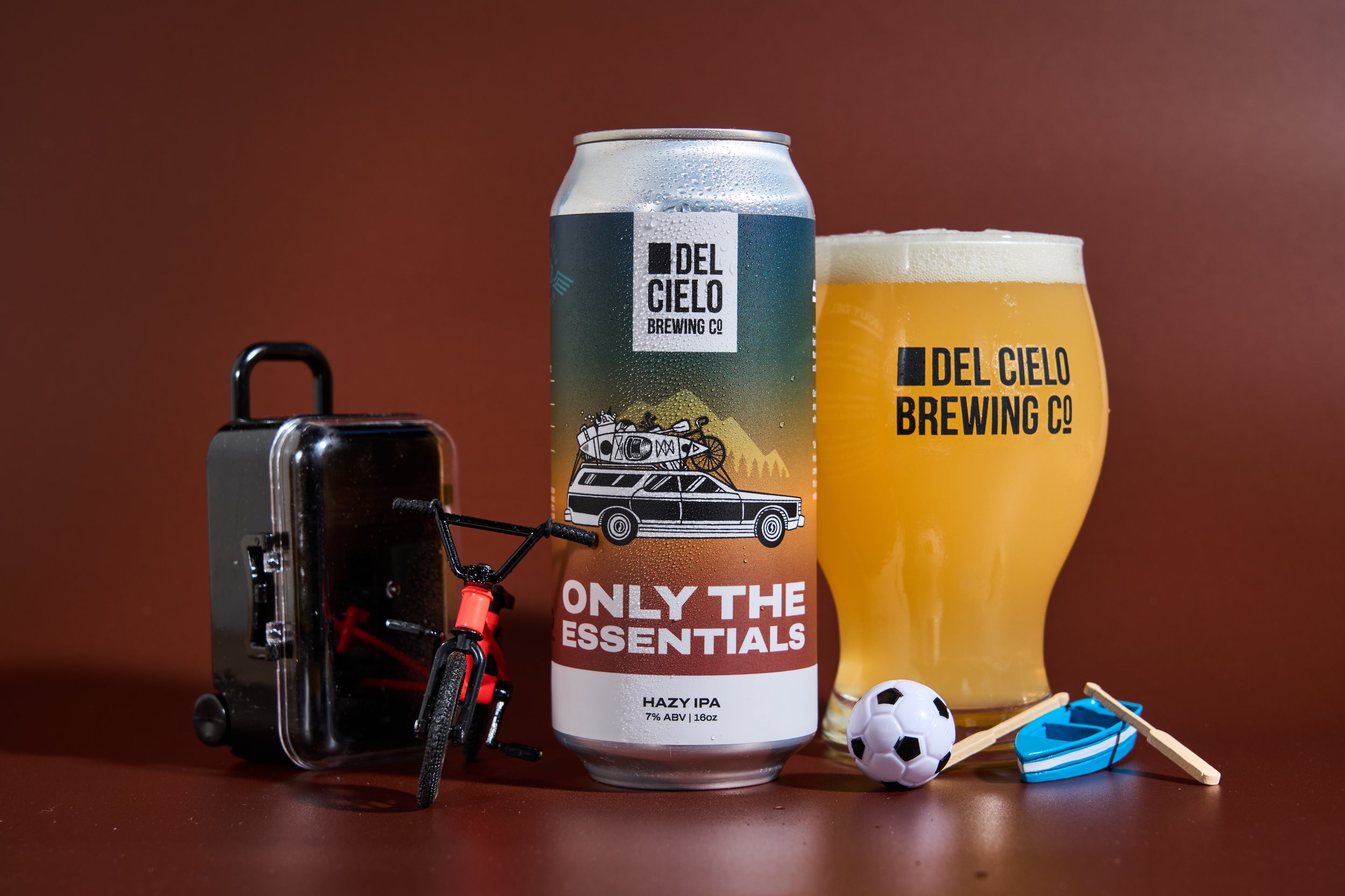 A can of Del Cielo Brewing Co hazy IPA beer, a glass of beer with foam, a small soccer ball, a toy boat, a small suitcase, and a miniature bicycle on a brown surface with a brown background.