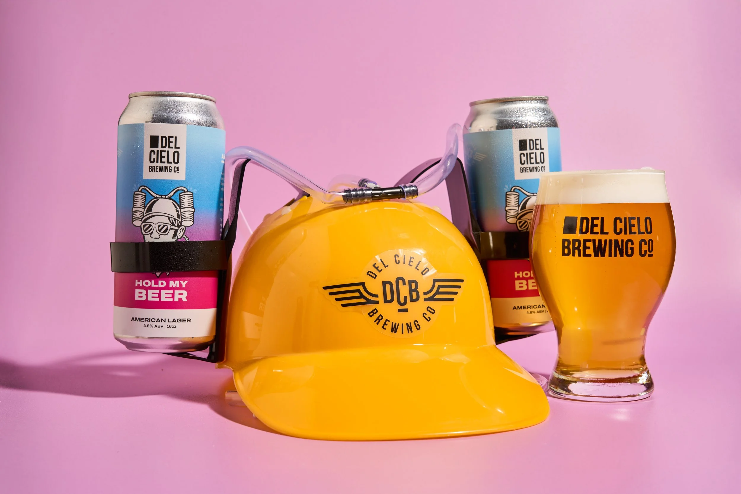 A collection of beer cans and a glass of beer from Del Cielo Brewing Co. against a pink background. The cans are labeled 'Hold My Beer' with an American lager style, and the glass shows a golden beer with a frothy head.