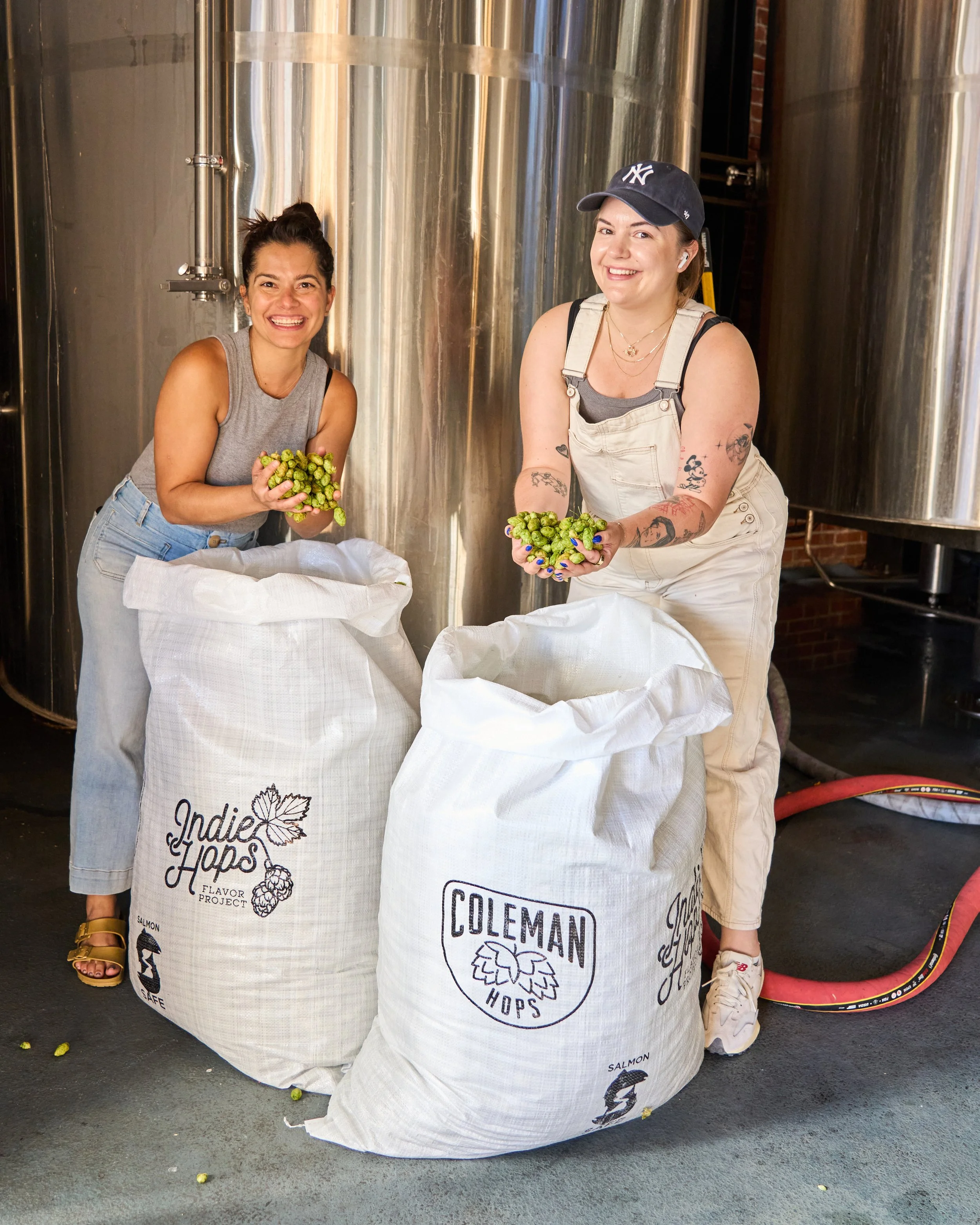 Two women holding freshly harvested hops standing behind large white sacks labeled "Indie Hops" and "Coleman Hops" in an industrial setting with large metal tanks in the background.