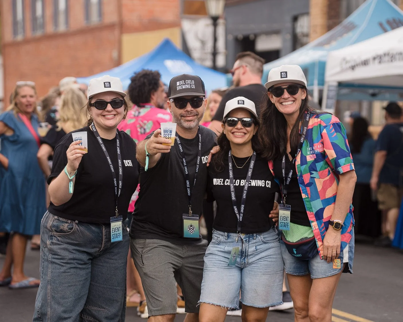 Four people smiling and holding small cups at an outdoor event, with tents and other attendees in the background.