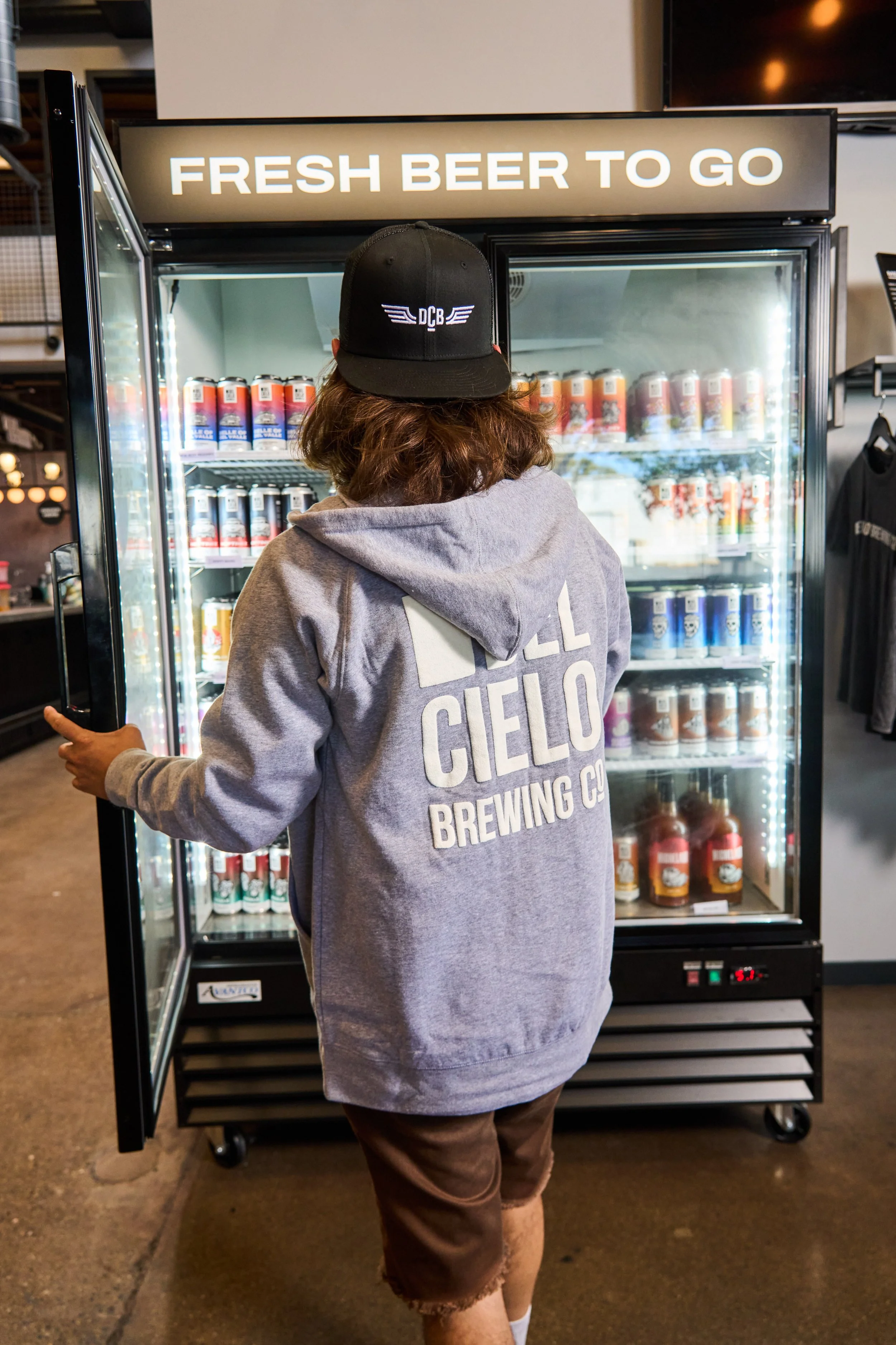 Person standing in front of a glass-door refrigerator with a sign that says 'FRESH BEER TO GO'. The person is wearing a black cap with a logo, a gray hoodie with writing on the back, and shorts. The refrigerator contains cans of beer.