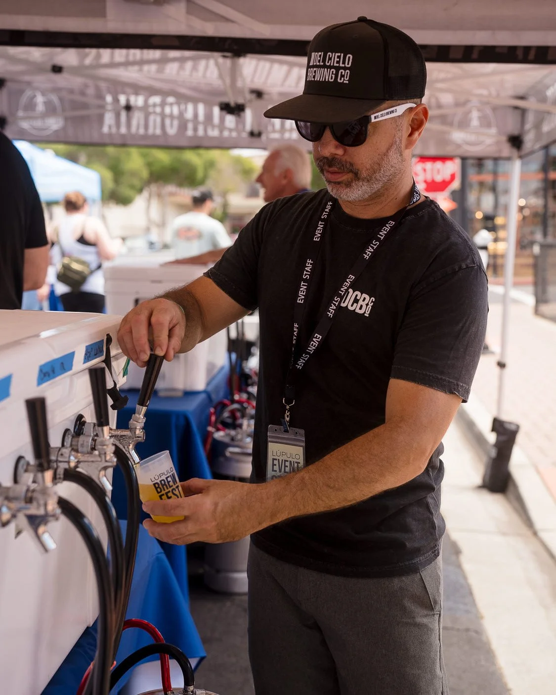 Man with gray beard, wearing black sunglasses, black cap, and black event staff shirt, pouring beer from a tap at an outdoor event.