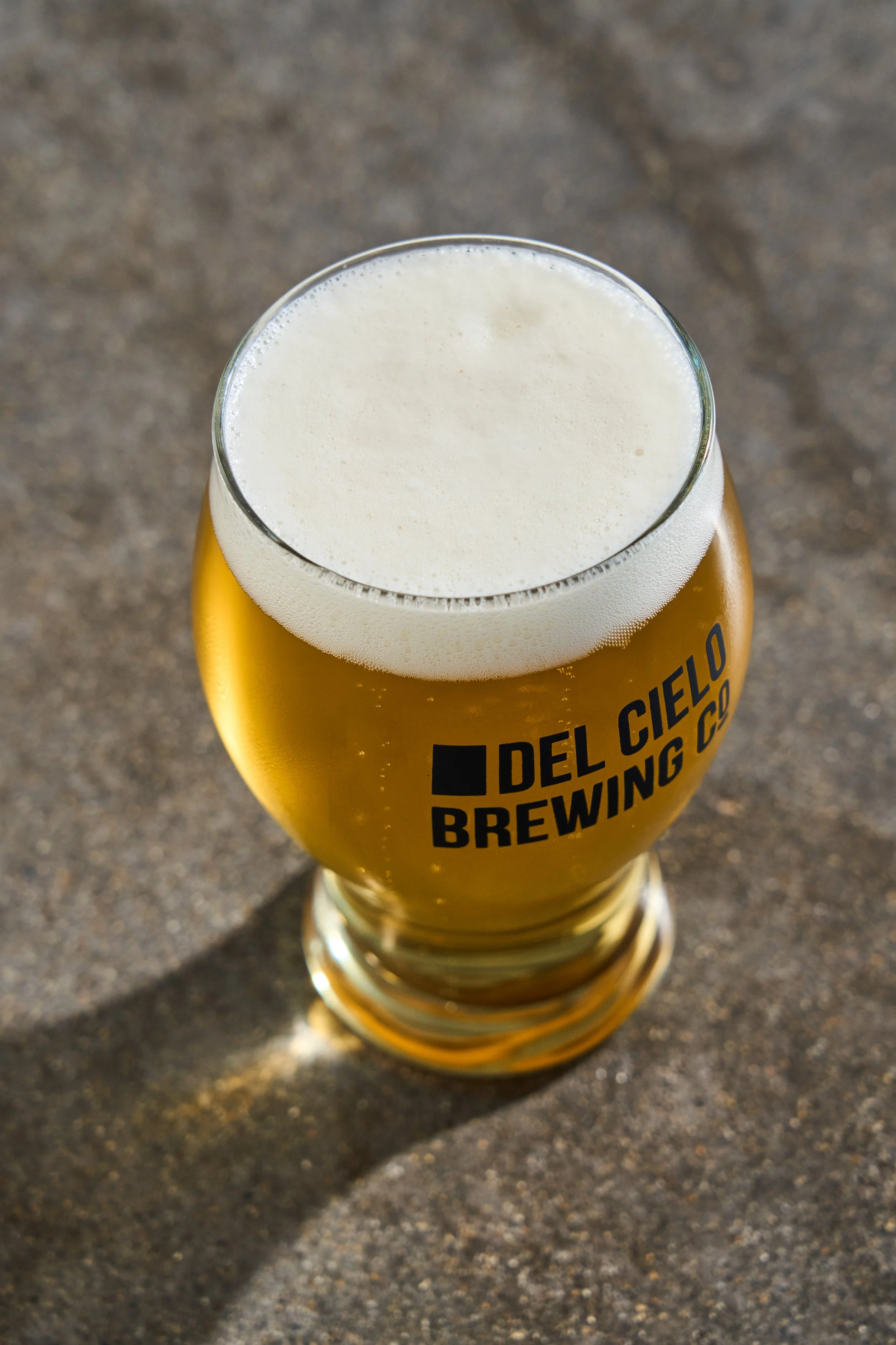 A glass of beer with a foamy head on a dark surface, with the text 'DEL CIELO BREWING CO' on the glass.