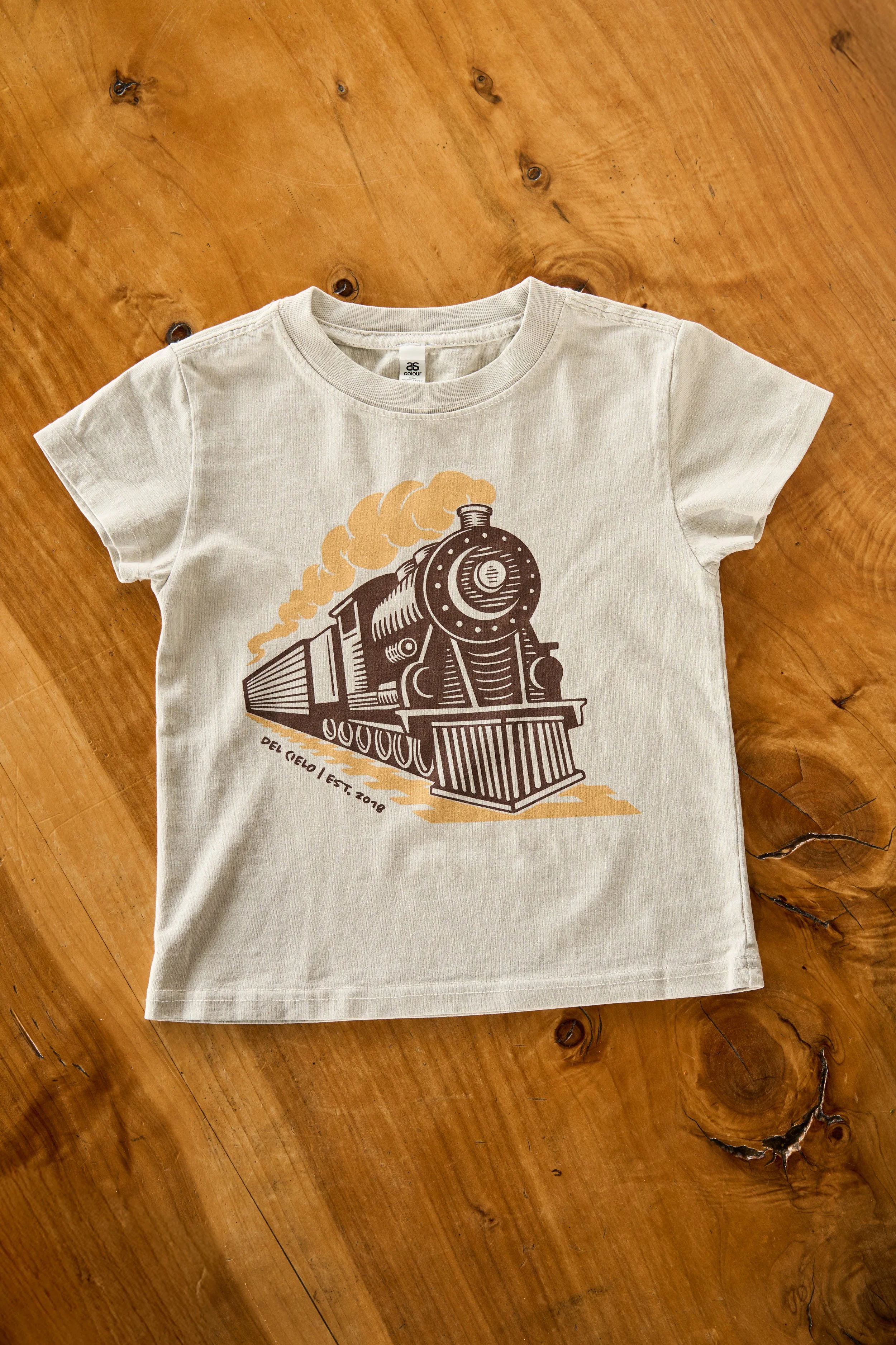 Beige T-shirt with a graphic of a steam train with smoke coming out of its chimney, on a wooden surface.