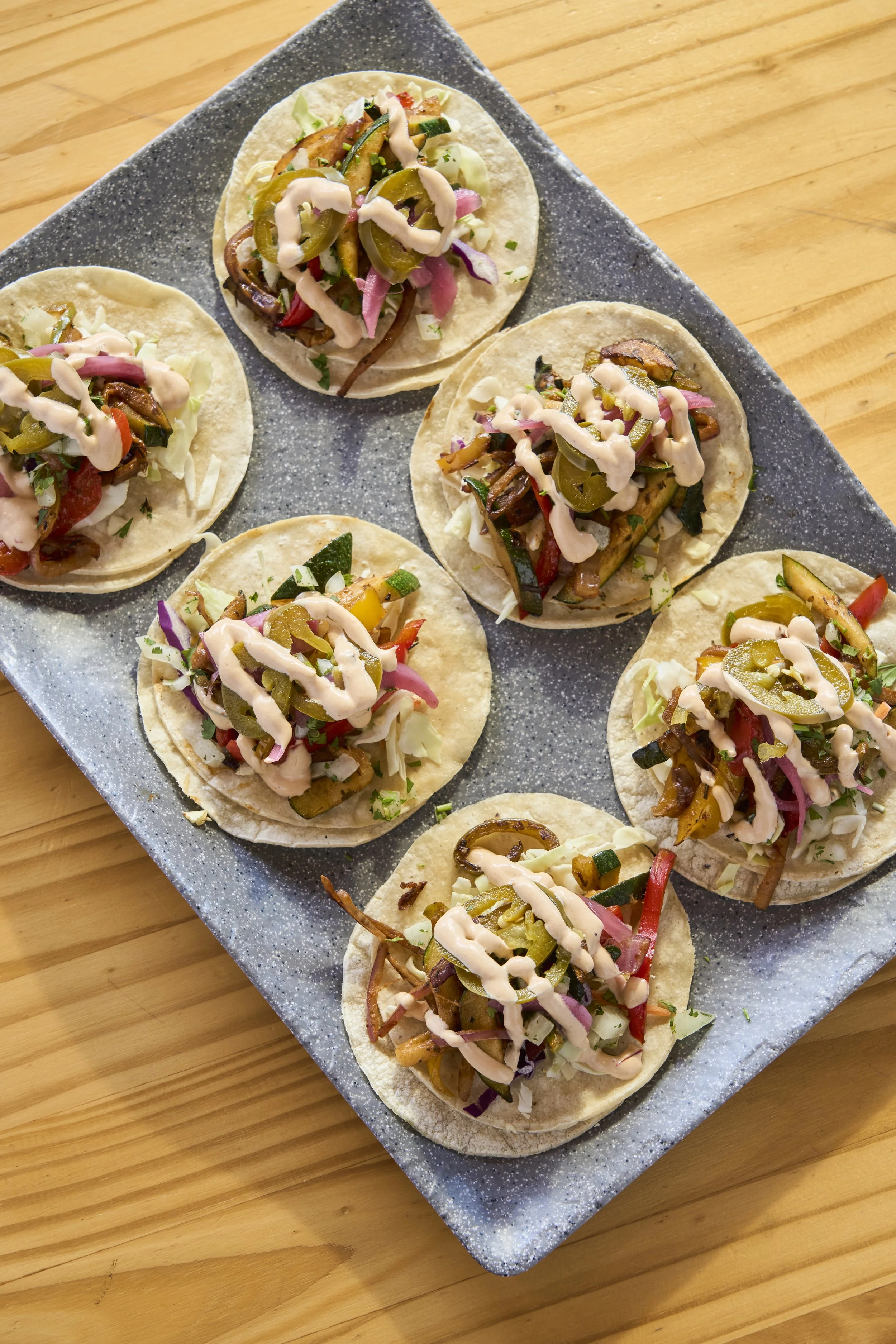 Close-up of several street-style tacos topped with shredded lettuce, pickled red onions, jalapeños, and drizzled with a creamy sauce on a black speckled rectangular plate on a wooden table.