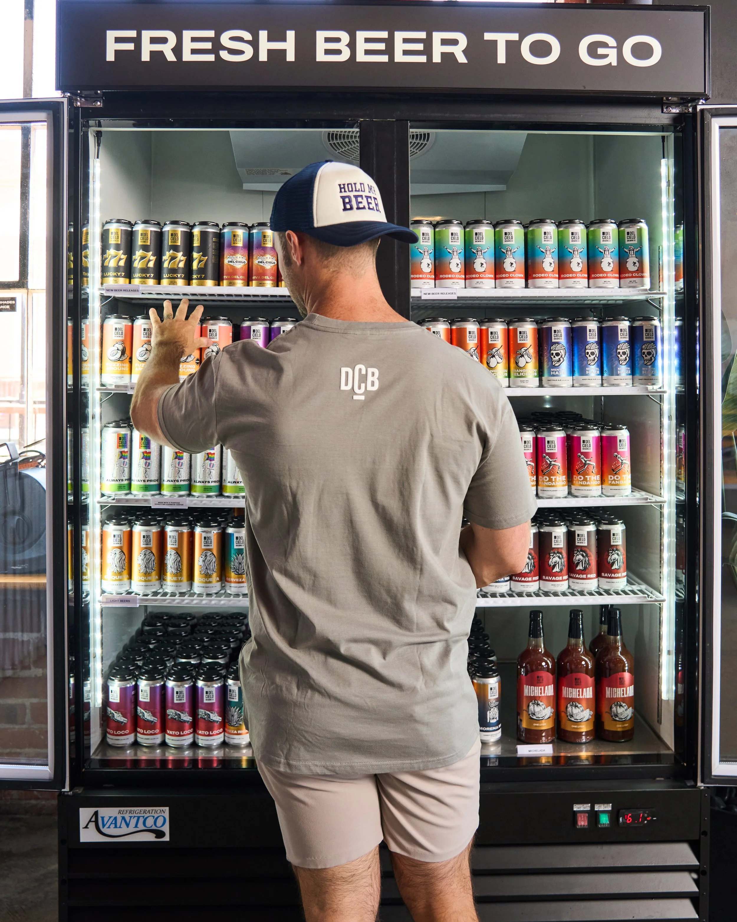 A man wearing a gray t-shirt and beige shorts is reaching for a can of beer from a refrigerated display case labeled "Fresh Beer To Go". The display case contains various cans of beer with colorful labels. The man is wearing a baseball cap that says 