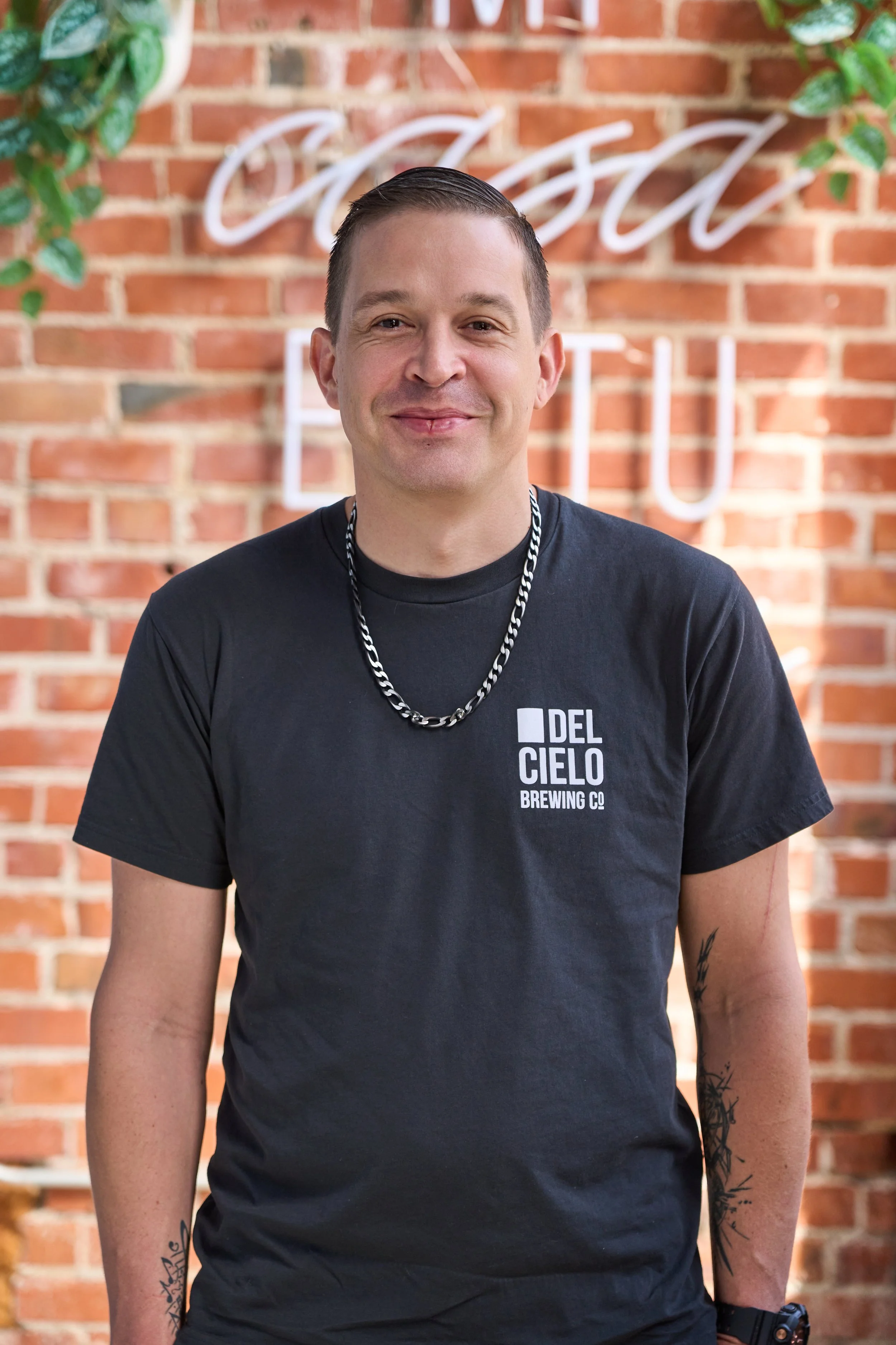 A smiling man with short dark hair wearing a black T-shirt with the 'Del Cielo Brewing Co.' logo, a silver chain necklace, and tattoos on arms, standing in front of a brick wall with a neon sign.