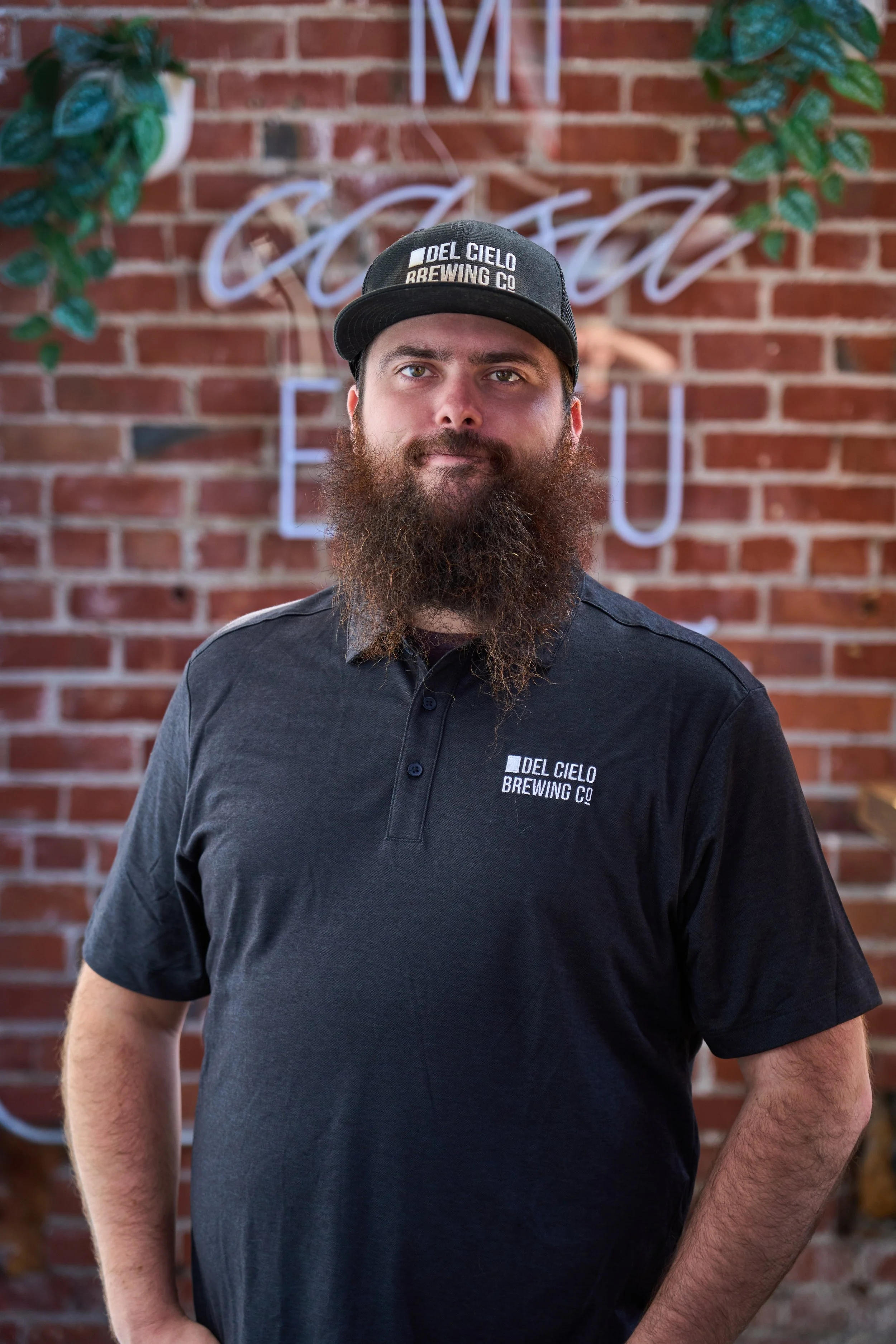 A bearded man wearing a black cap and a black polo shirt with the logo 'Del Cielo Brewing Co' standing in front of a brick wall with the word 'Cielo' in illuminated neon letters and green plants on each side.