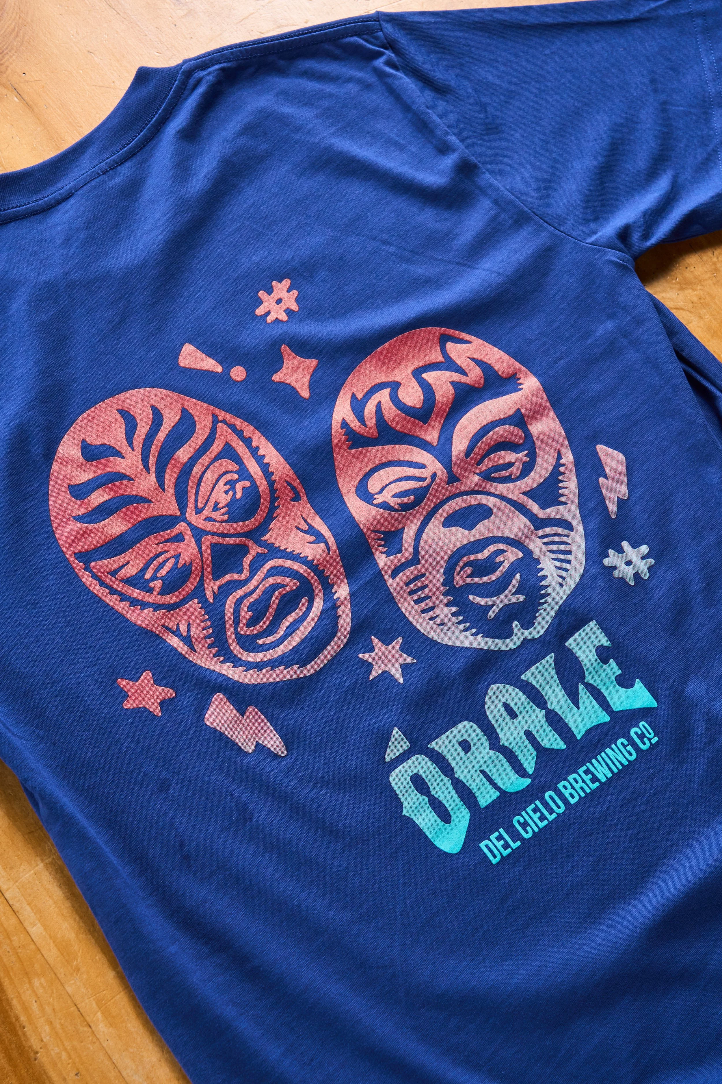 Blue T-shirt with a logo featuring lucha libre masks and stars, along with the text 'Orale', and additional text 'Del Cielo Brewing Co.', lying on a wooden surface.