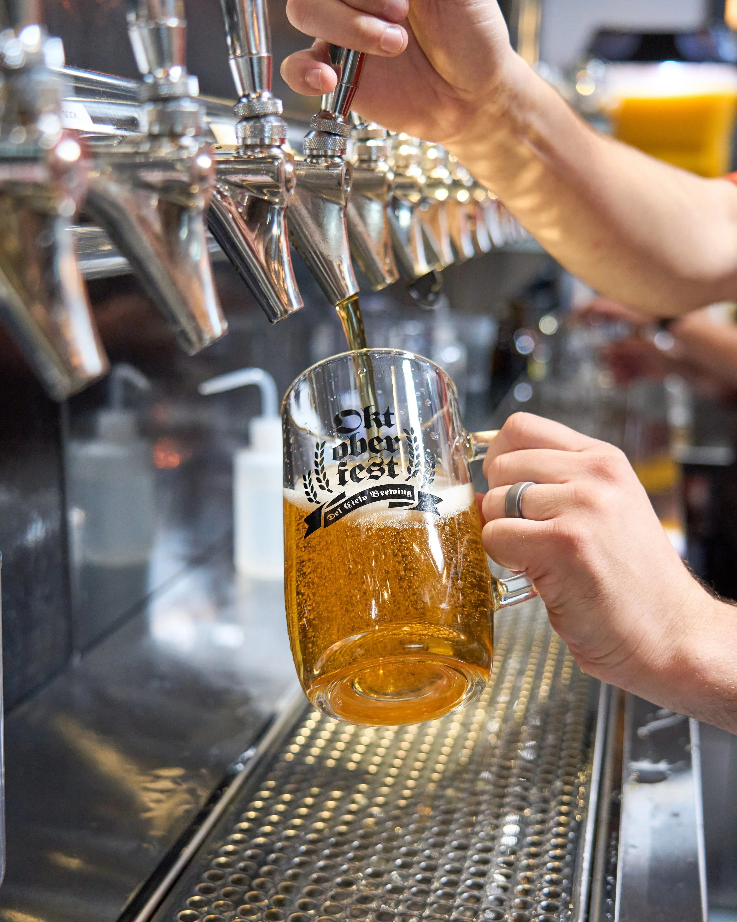 A person is pouring beer from a tap into a glass at a brewery or bar. The glass has a logo that says 'oktoberfest' with decorative laurel leaves around it.